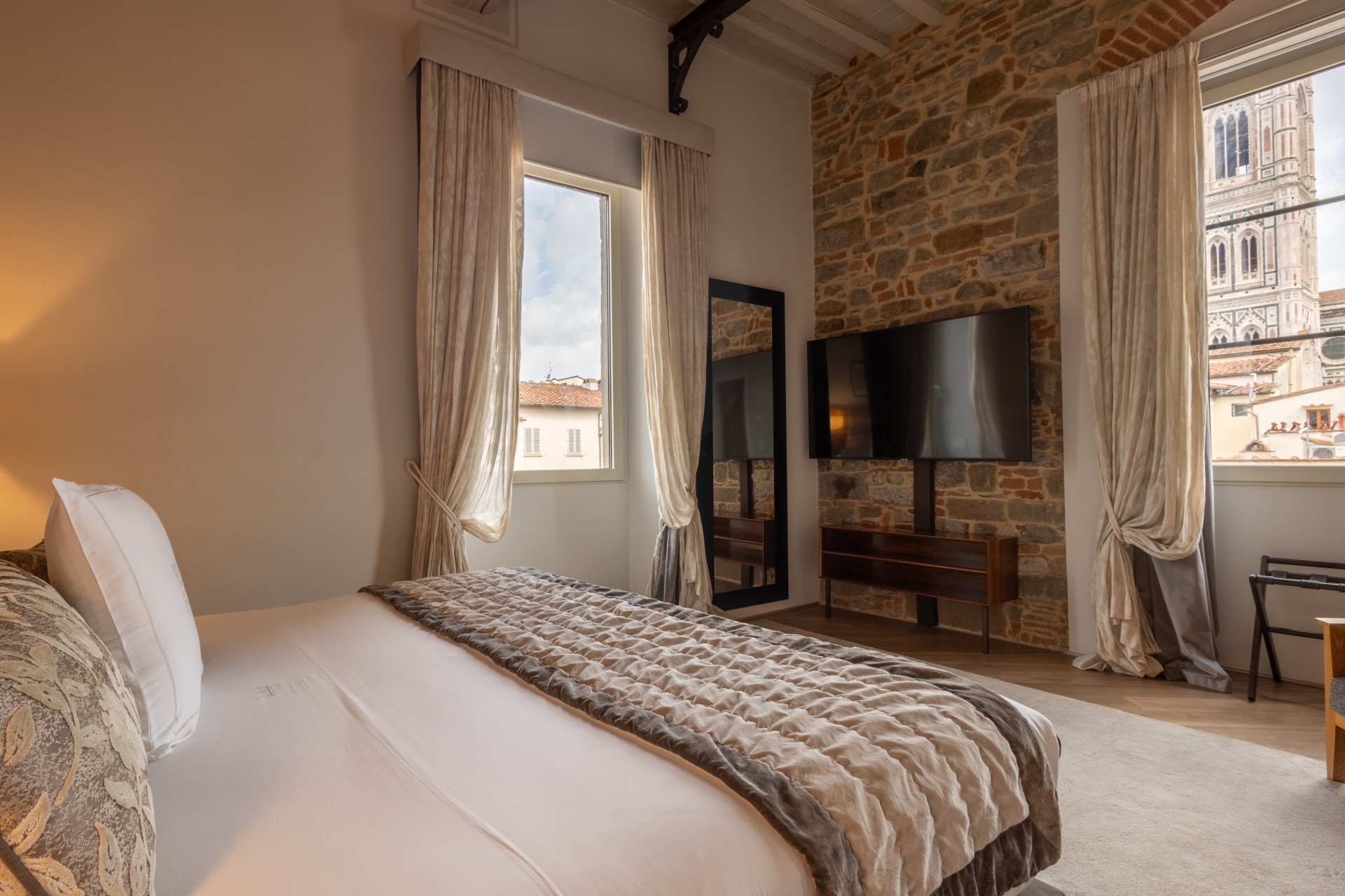 Side-angle view of a luxury hotel suite bedroom showing a king-size bed with plush throw pillows, exposed stone wall with a mounted flat-screen TV, and windows offering views of Giotto's Bell Tower.