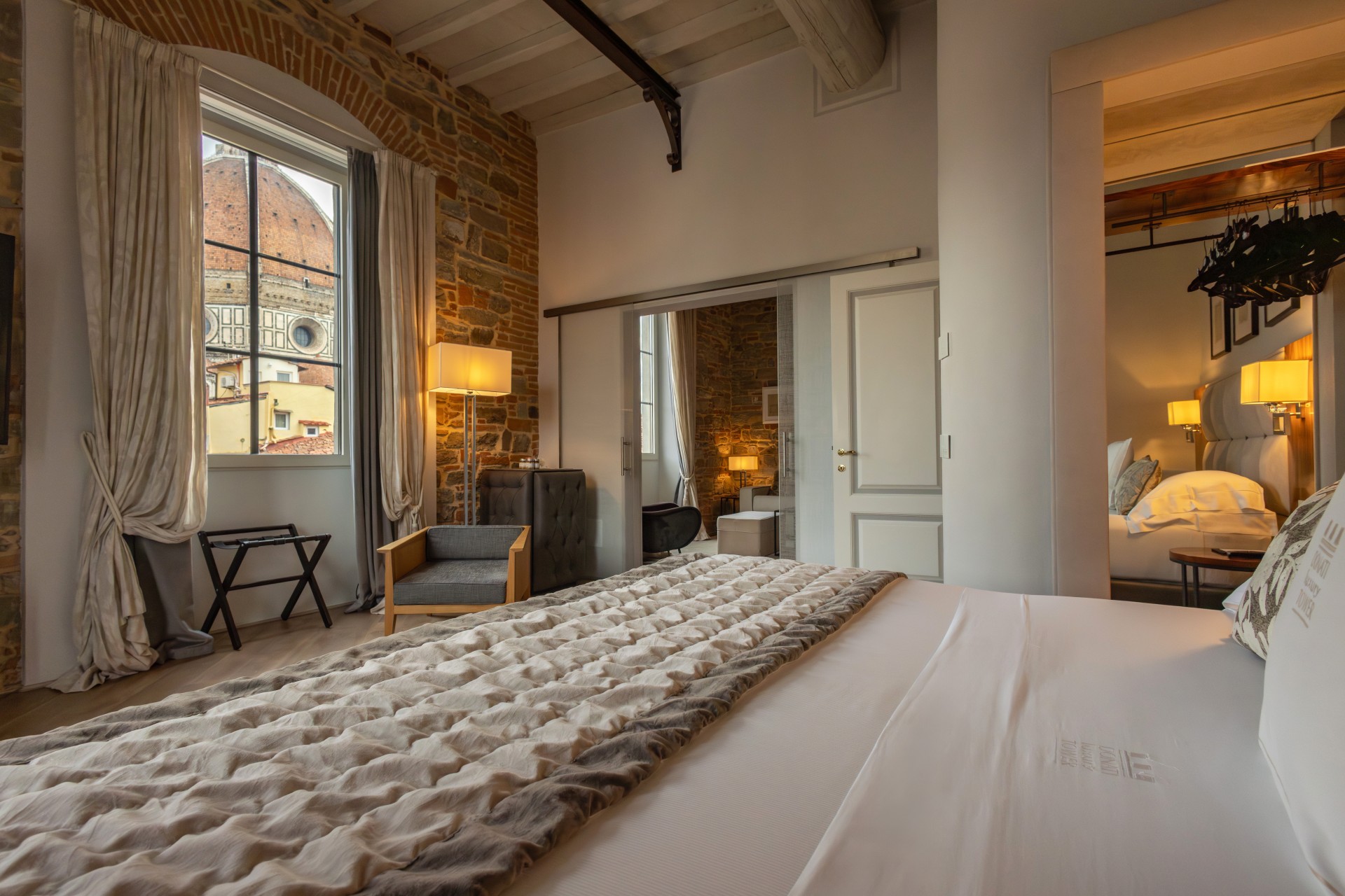 Interior view of a luxury hotel suite looking across a king-size bed toward a window framing Florence Cathedral's dome, with exposed brick and wood-beamed ceilings.