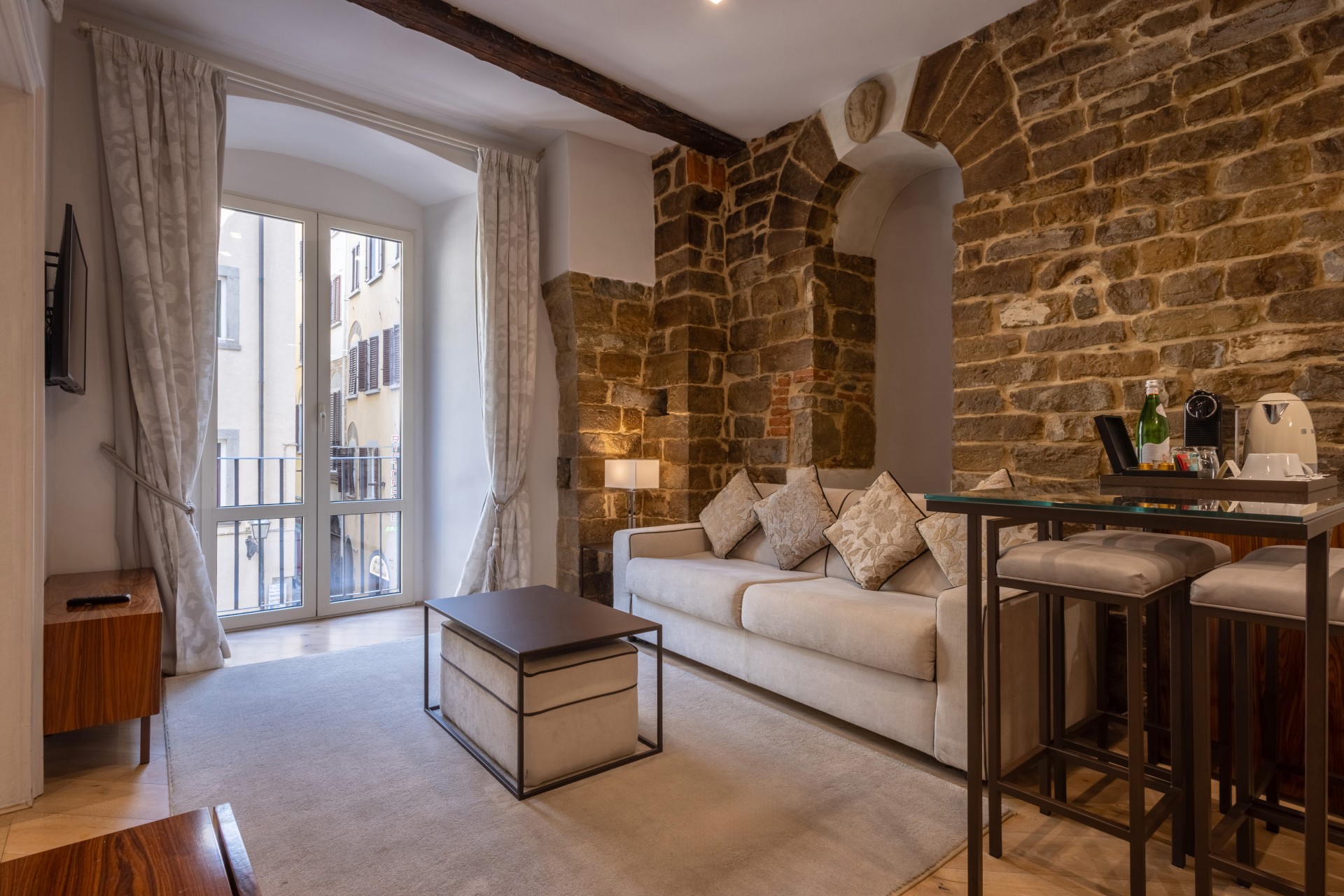 Hotel suite sitting room with exposed stone accent walls, arched architectural details, a light beige couch with patterned throw pillows, a coffee table with an ottoman, glass balcony doors, and a high-top glass table with bar stools.