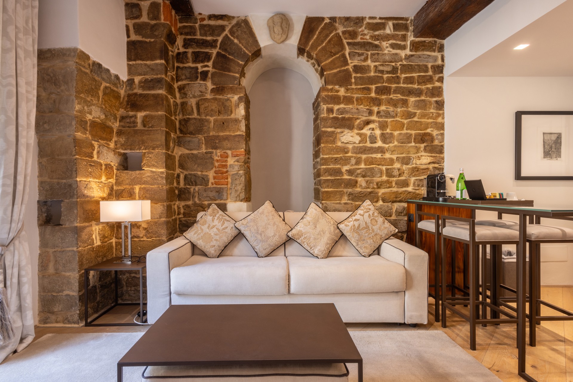 Elegant hotel sitting room featuring an exposed stone archway wall, a light cream sofa with patterned throw pillows, a dark coffee table, and a high-top bar table with stools.