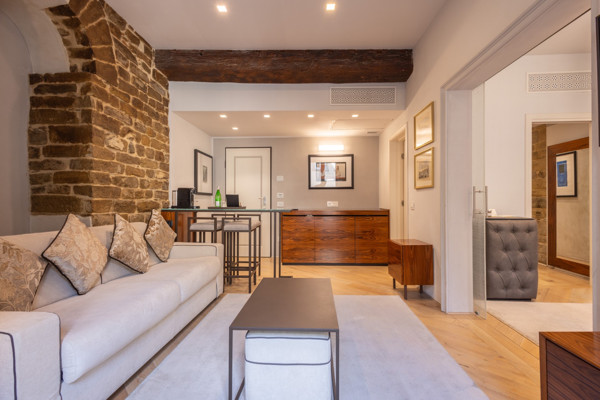 Spacious suite living area featuring a plush white sofa, dark coffee table on a light rug, rustic stone column, dark wooden beam ceiling, and a high-top bar counter.