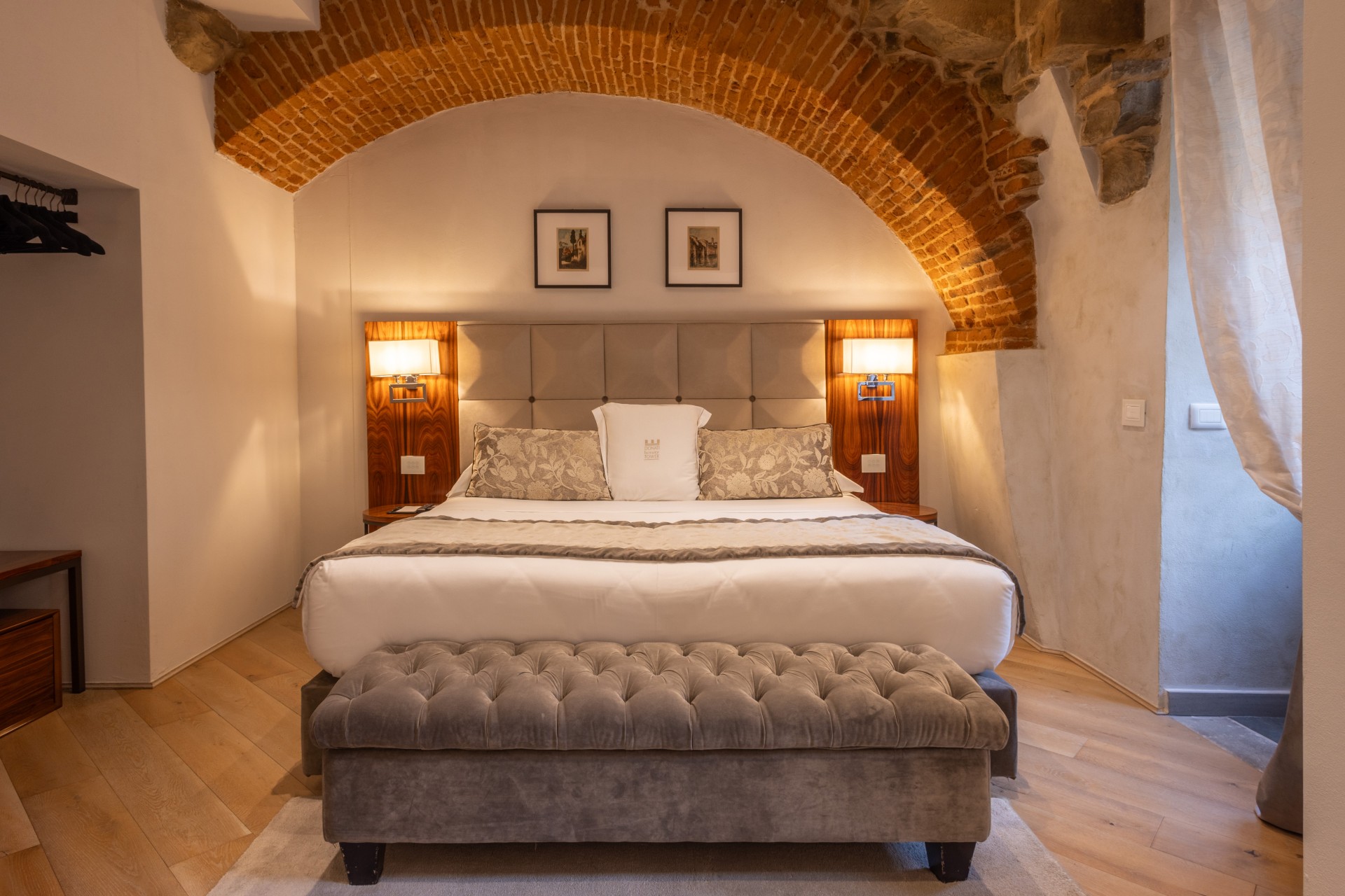 Luxurious hotel bedroom with a large bed beneath an exposed red-brick arch, framed by warm wood wall-mounted sconces, a tufted gray ottoman at the foot of the bed, and light hardwood flooring.