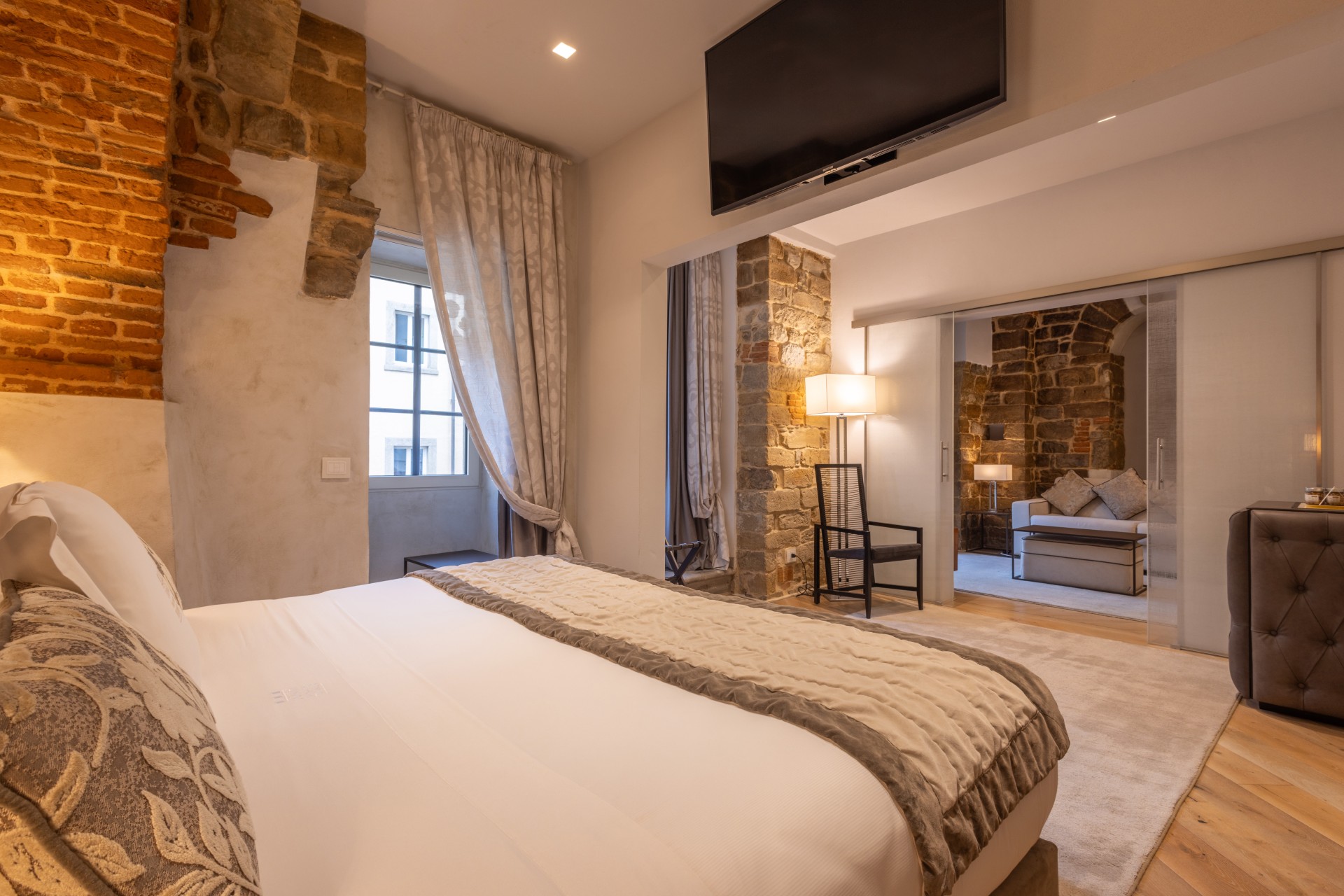 Hotel suite bedroom with a comfortable bed in the foreground, exposed brick wall accents, a wall-mounted TV, and glass sliding doors opening into an adjoining stone-walled living area.