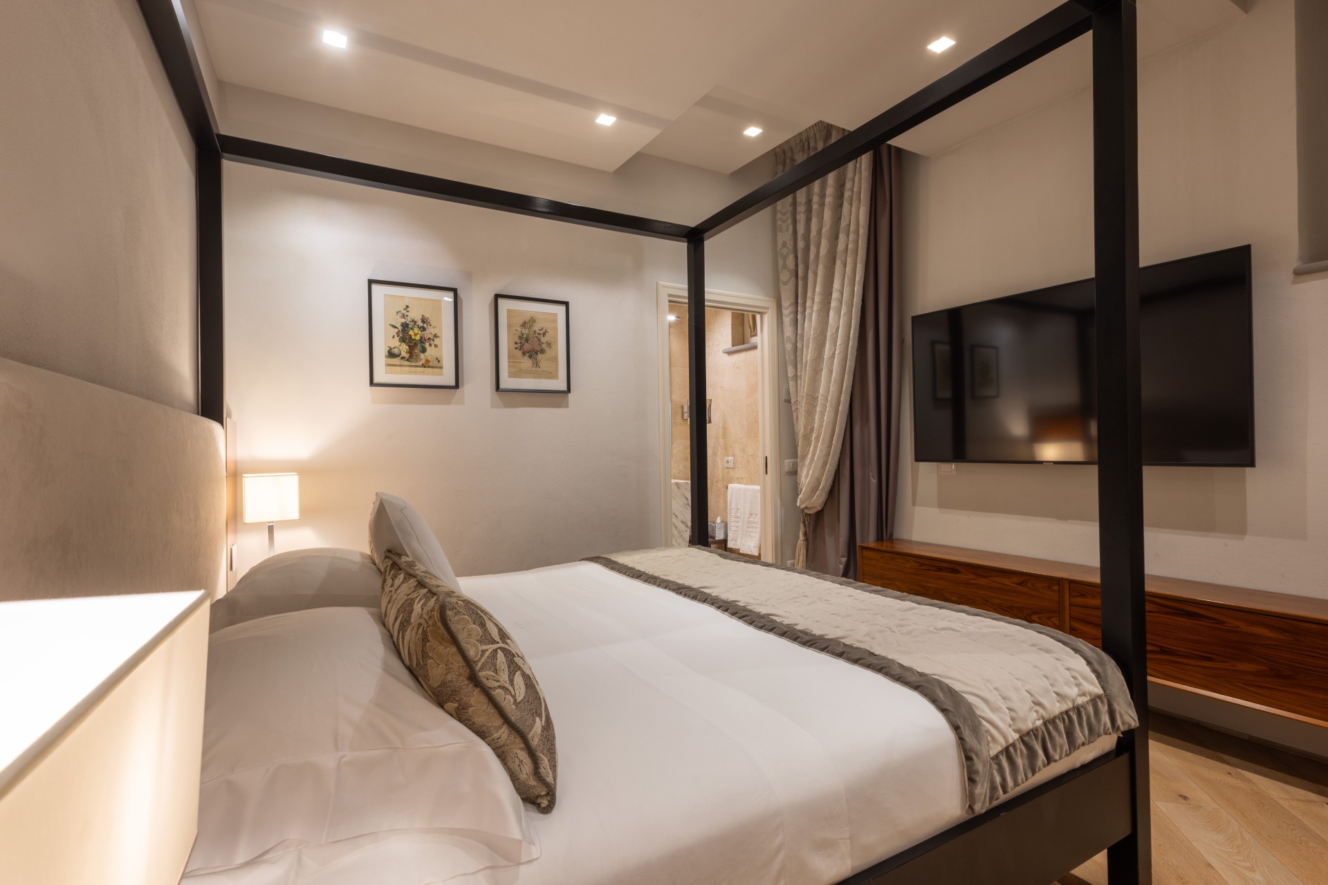 Modern hotel bedroom with a black four-poster canopy bed, white bedding, framed botanical prints, wall-mounted flat-screen TV, and an open door leading to an en-suite bathroom.