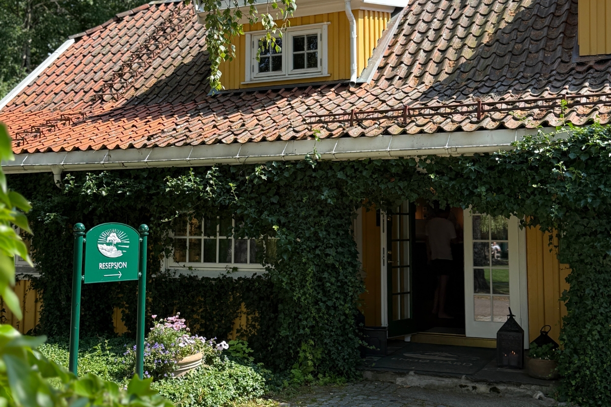 A charming yellow wooden cottage with a terracotta tile roof, ivy climbing around the entrance, potted flowers out front, and a green directional sign pointing to the reception ('Resepsjon').
