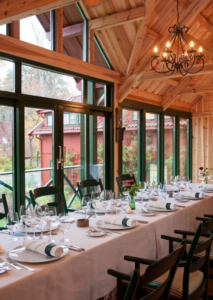A rustic dining room with high vaulted wooden panel ceilings, dark metal wrought-iron chandeliers, and floor-to-ceiling glass windows framing red wooden buildings outside. A long table dressed with a white tablecloth, formal place settings, crystal glassw