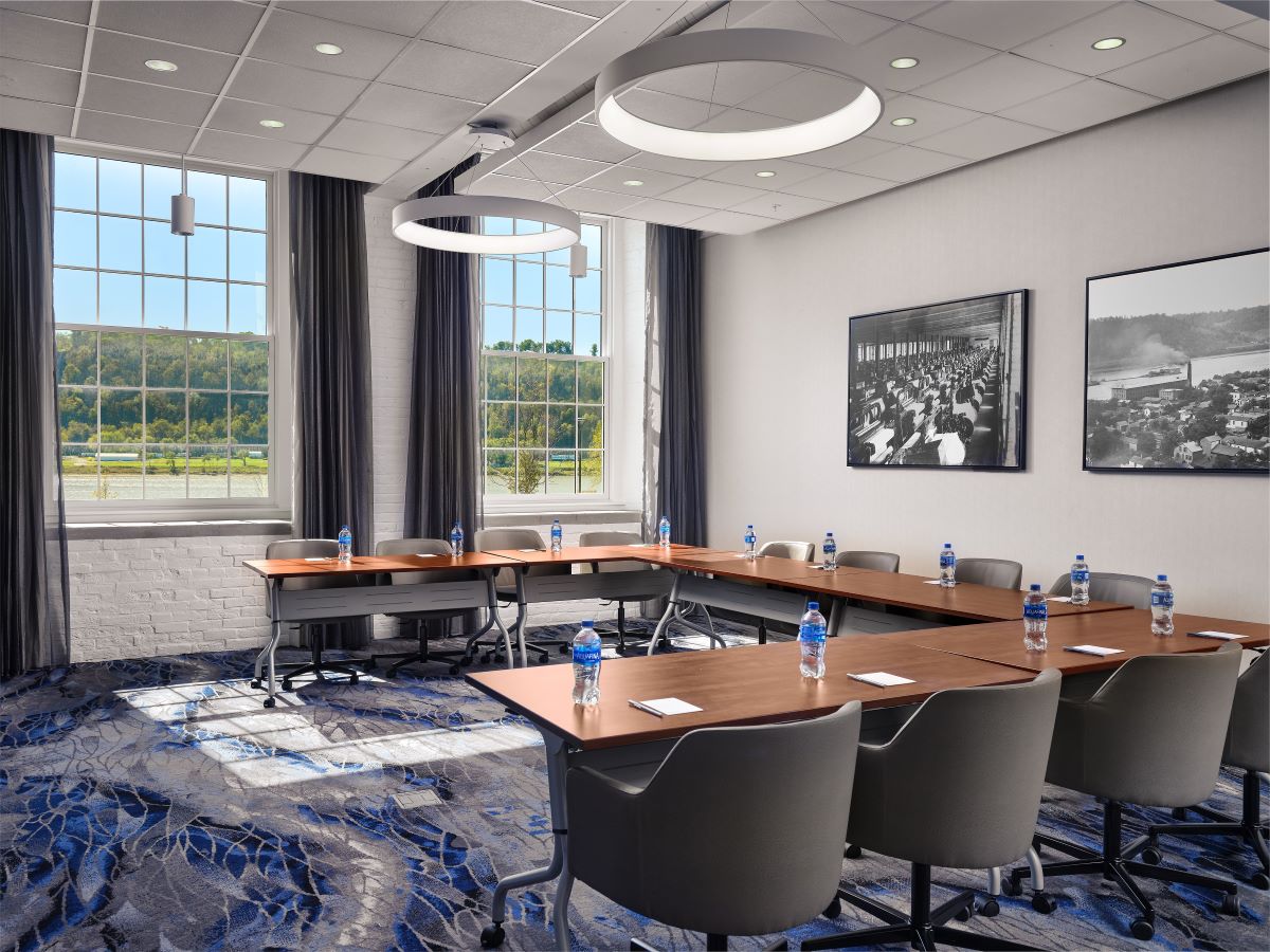 Modern conference room with U-shaped table setup, grey rolling chairs, ring halo light fixtures, large windows overlooking a scenic river, and black-and-white photos on the wall.