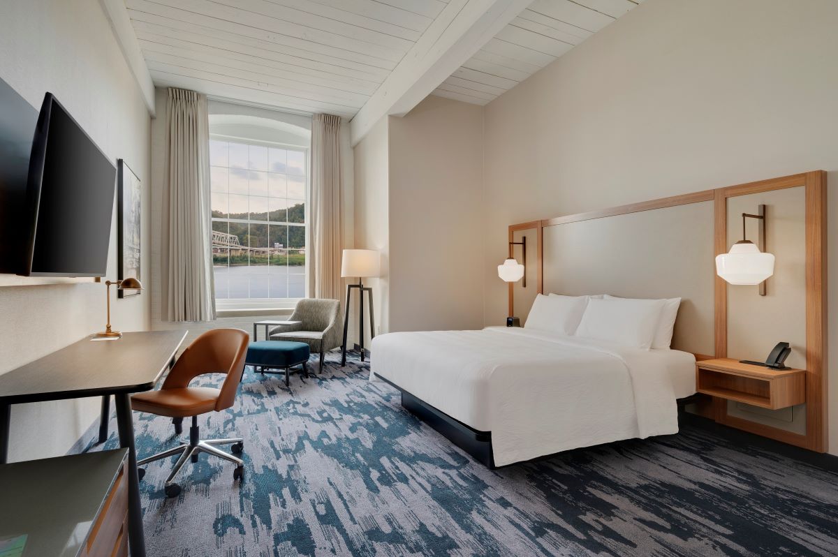 Modern hotel room with high exposed beam ceilings, a king bed with an upholstered headboard, a dark wood desk with a cognac leather office chair, and an arched window overlooking a river view.