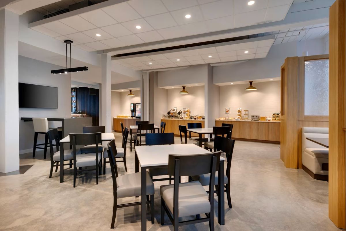 Modern hotel breakfast dining area featuring polished concrete floors, light wood serving buffets, and square tables surrounded by dark-framed chairs with grey cushions.