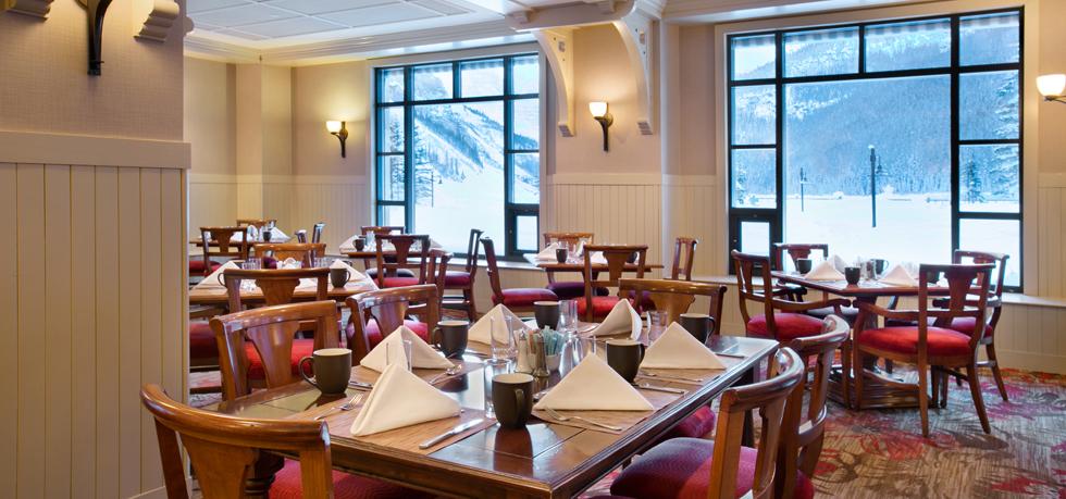 A cozy, well-lit dining area featuring wooden tables set with neatly folded white napkins and mugs, paired with wooden chairs with red seats. Large grid windows offer a scenic view of a snowy mountain landscape outdoors.