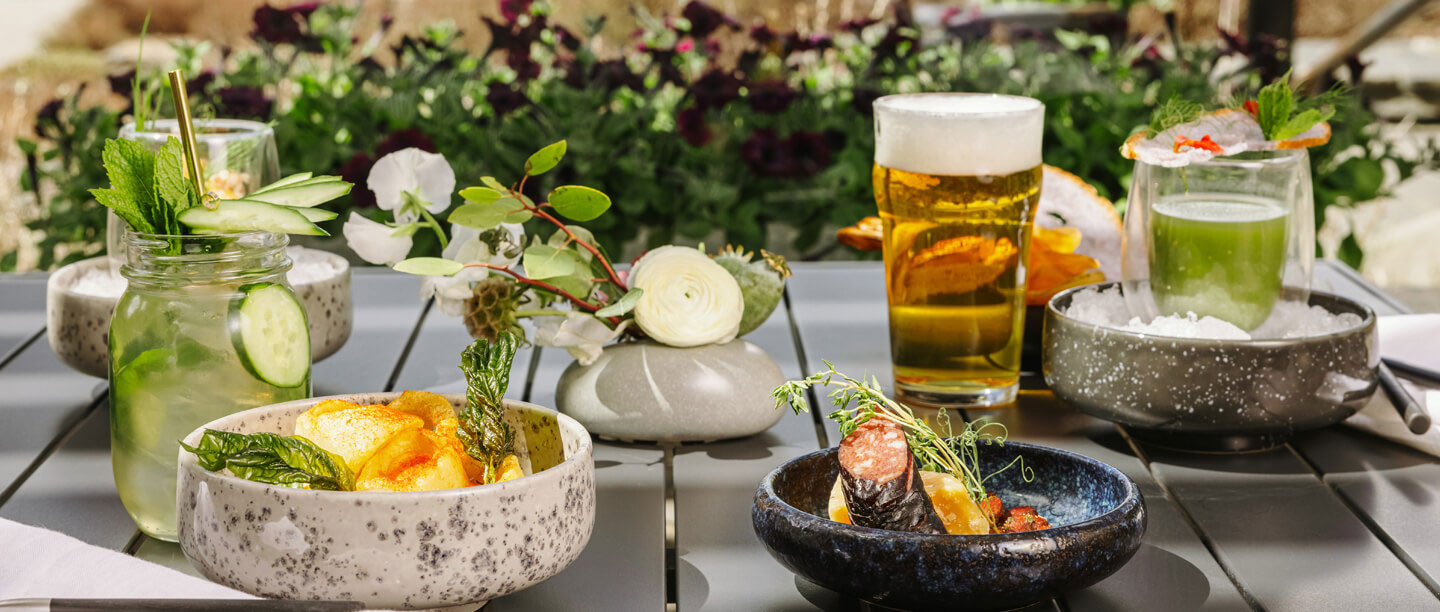 An outdoor dining setup on a slatted grey table featuring various drinks and appetizers. Items include a glass jar cocktail garnished with mint and cucumber slices, a bowl of crispy potato chips, a glass of foamy beer, a dark ceramic bowl with a gourmet s