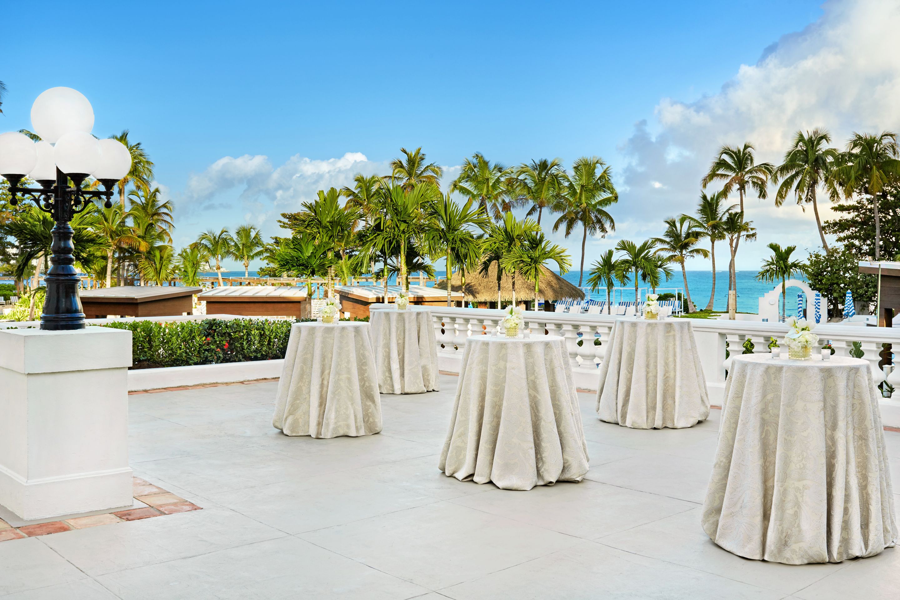 a group of tables with white cloths on a patio with palm trees and a body of water