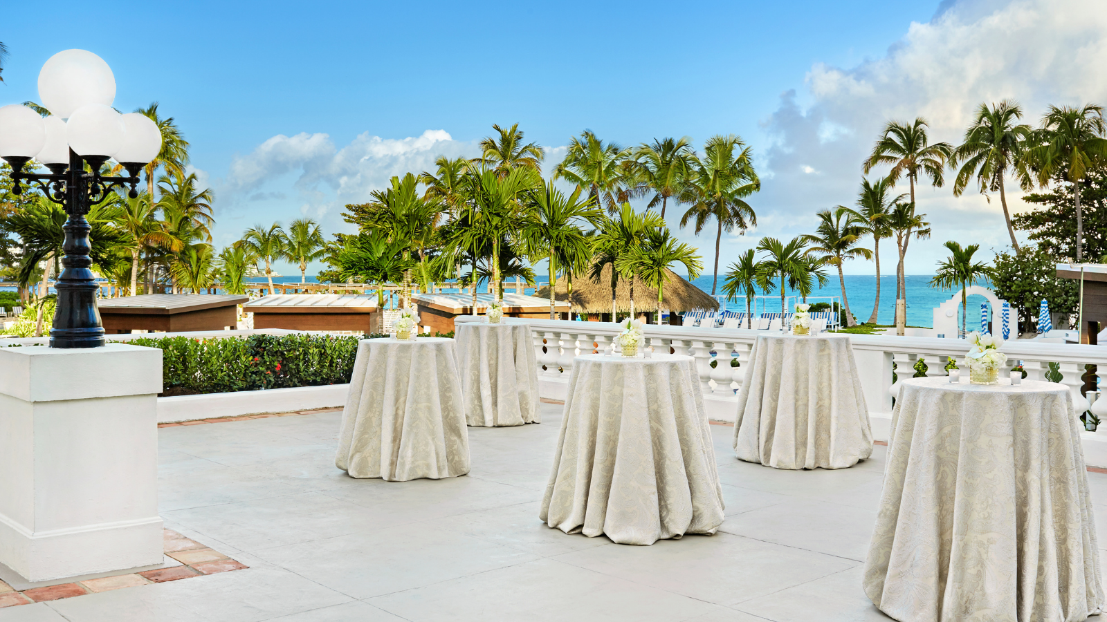 a group of tables with white cloths on a patio with palm trees and a beach