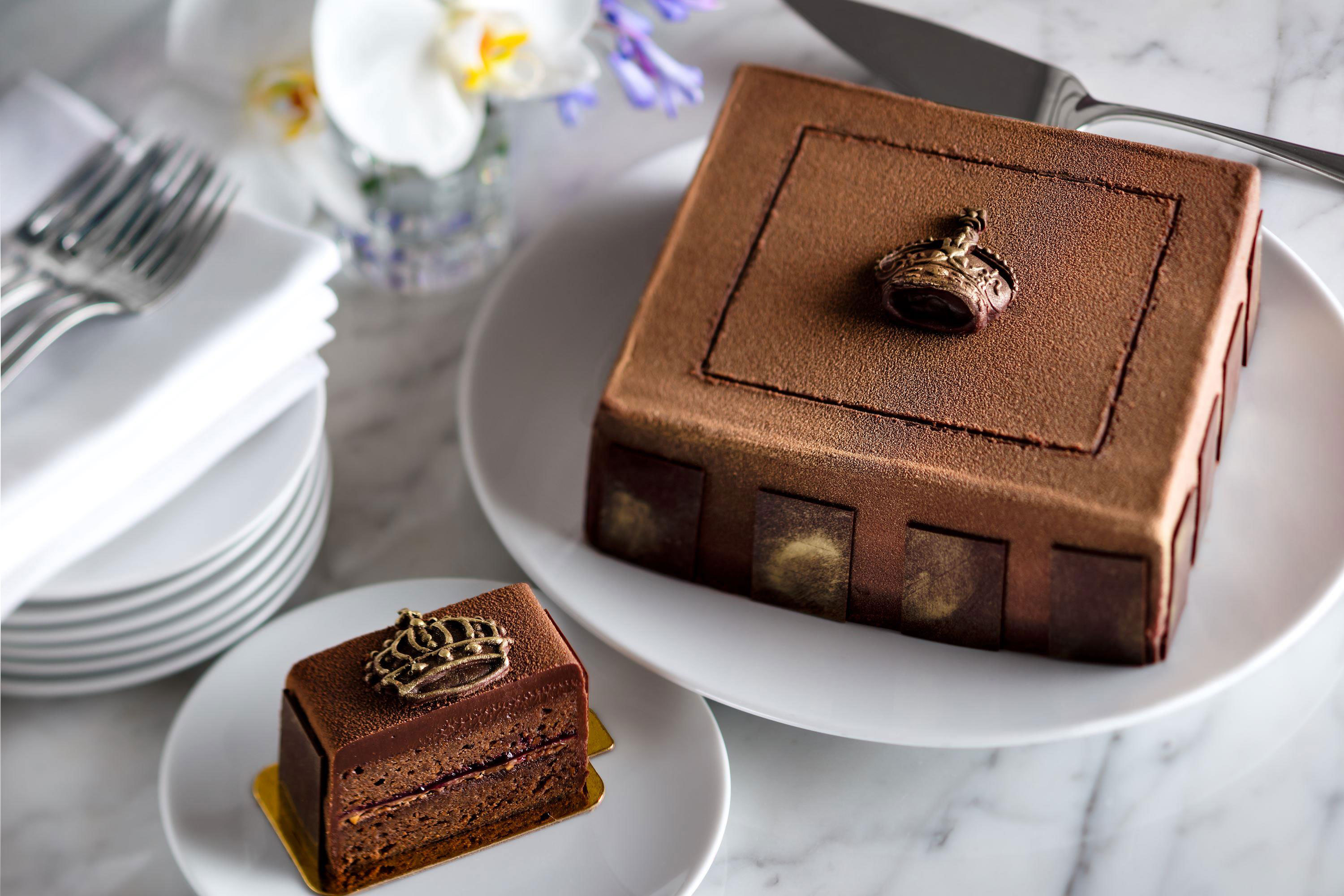 Decadent square chocolate cake dusted with cocoa powder and topped with a metallic gold crown insignia, next to an individual slice showing layered chocolate and fruit filling.