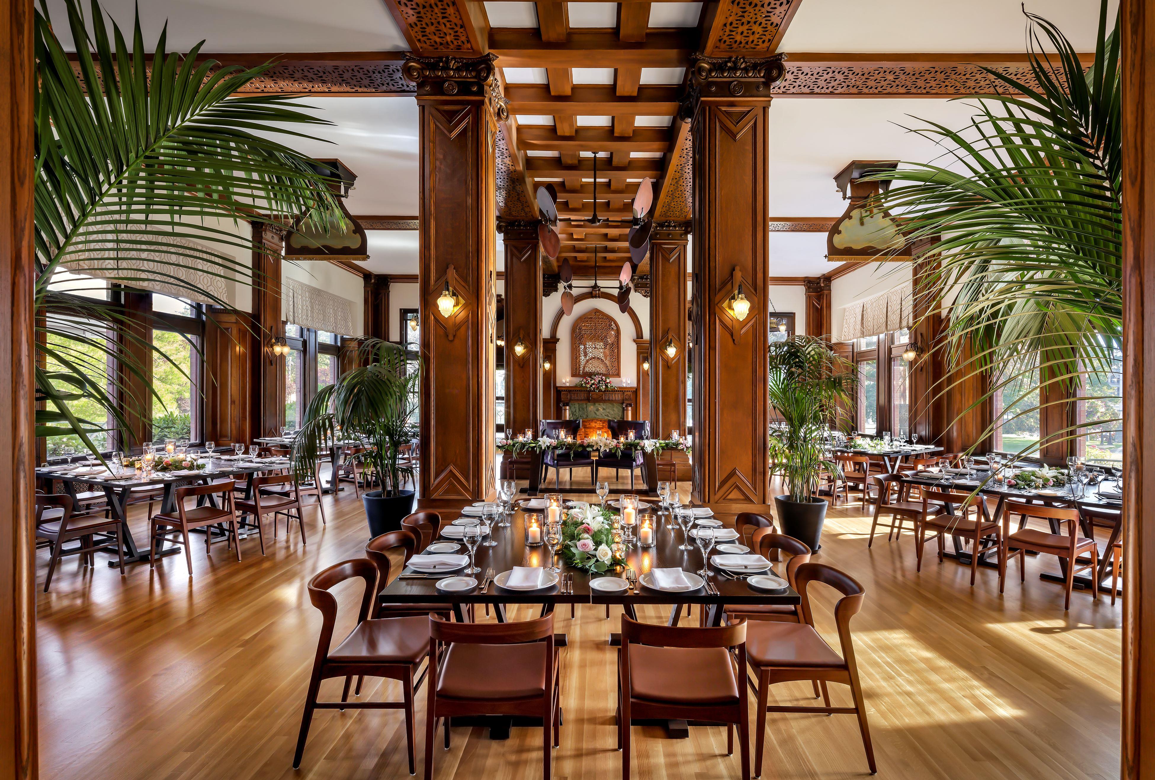 Grand dining room with rich wood columns, coffered ceilings, potted palm trees, and long wooden dining tables set for an event with candles and floral arrangements.