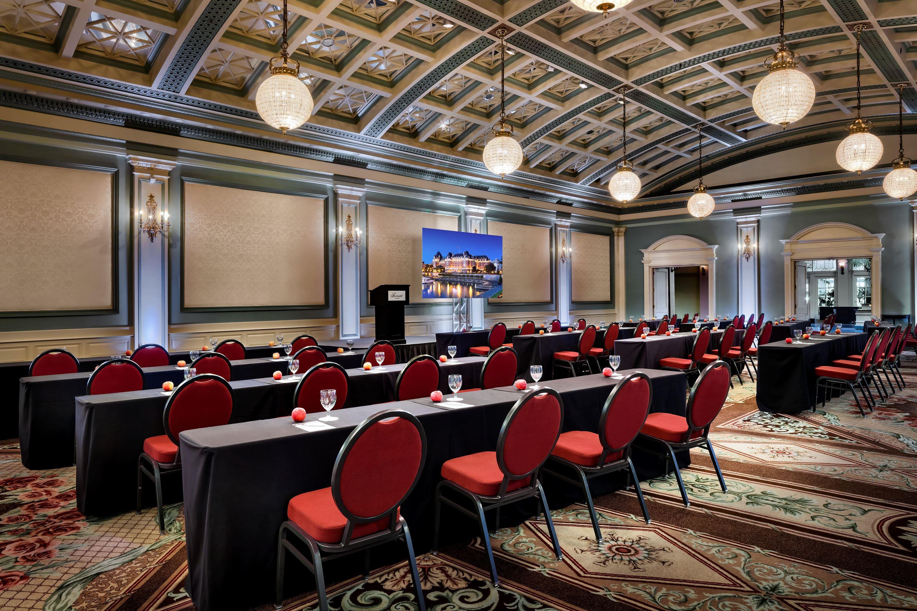 Large conference room set up classroom-style with long black-clothed tables, red padded chairs, globe pendant lights, an ornate patterned ceiling, and a presentation screen on stage.