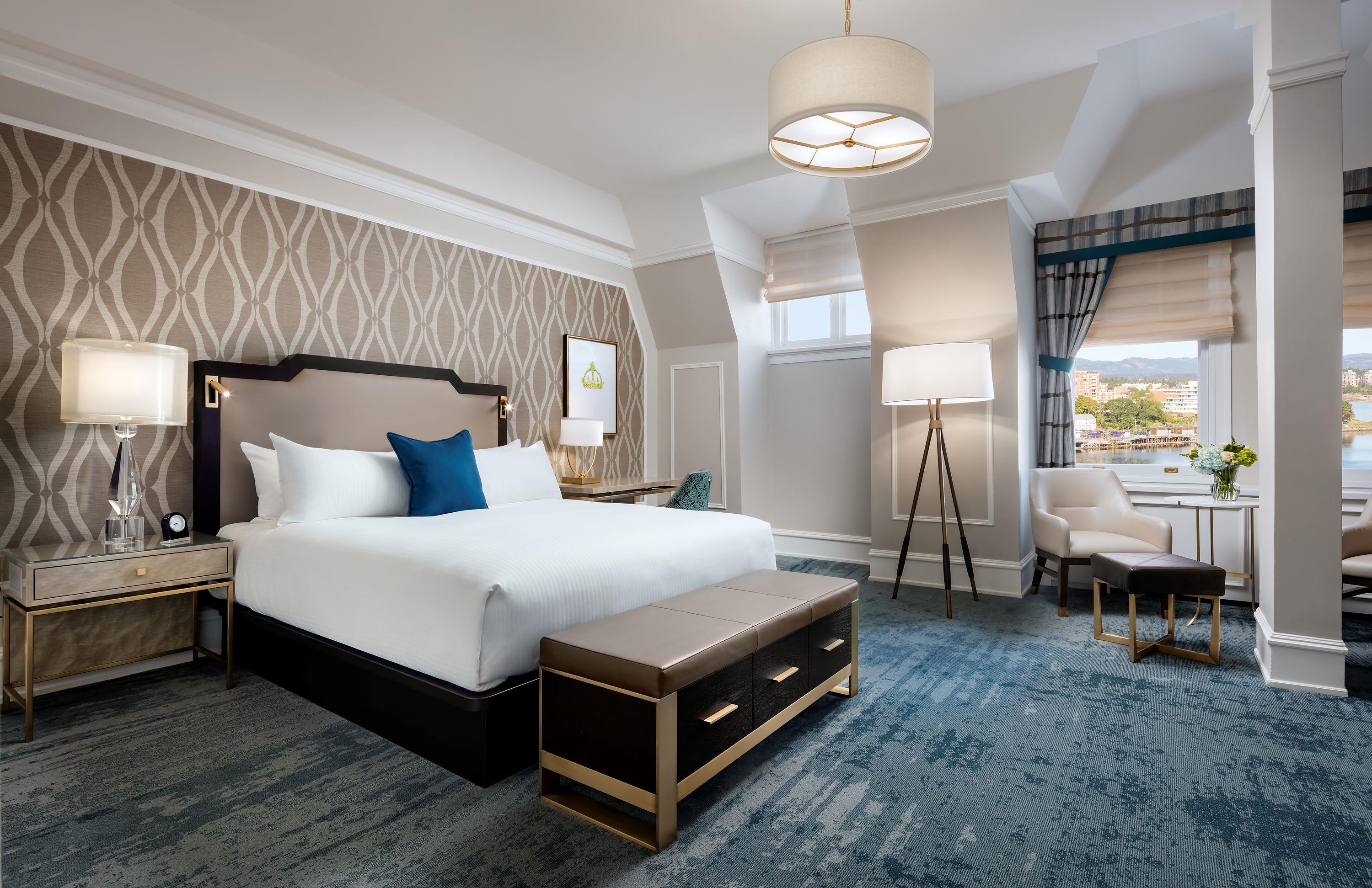 Spacious luxury hotel bedroom with a king bed, patterned feature wallpaper, blue textured carpet, a leather bench at the foot of the bed, and a seating area by windows with water views.