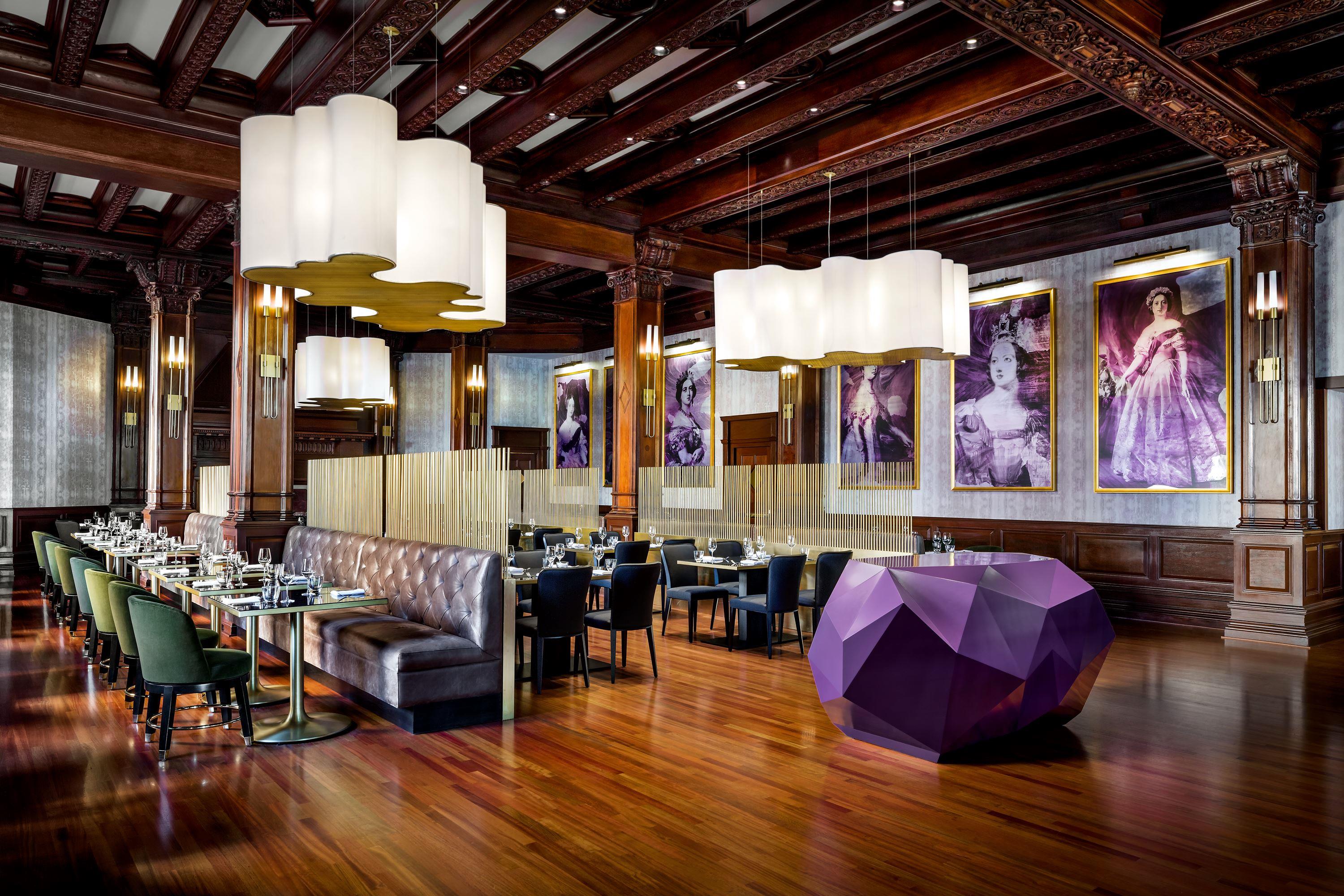 Upscale restaurant featuring dark wood ceiling beams and columns, large cloud-like pendant lights, a geometric purple reception counter, and large purple-toned portraits along the back wall.