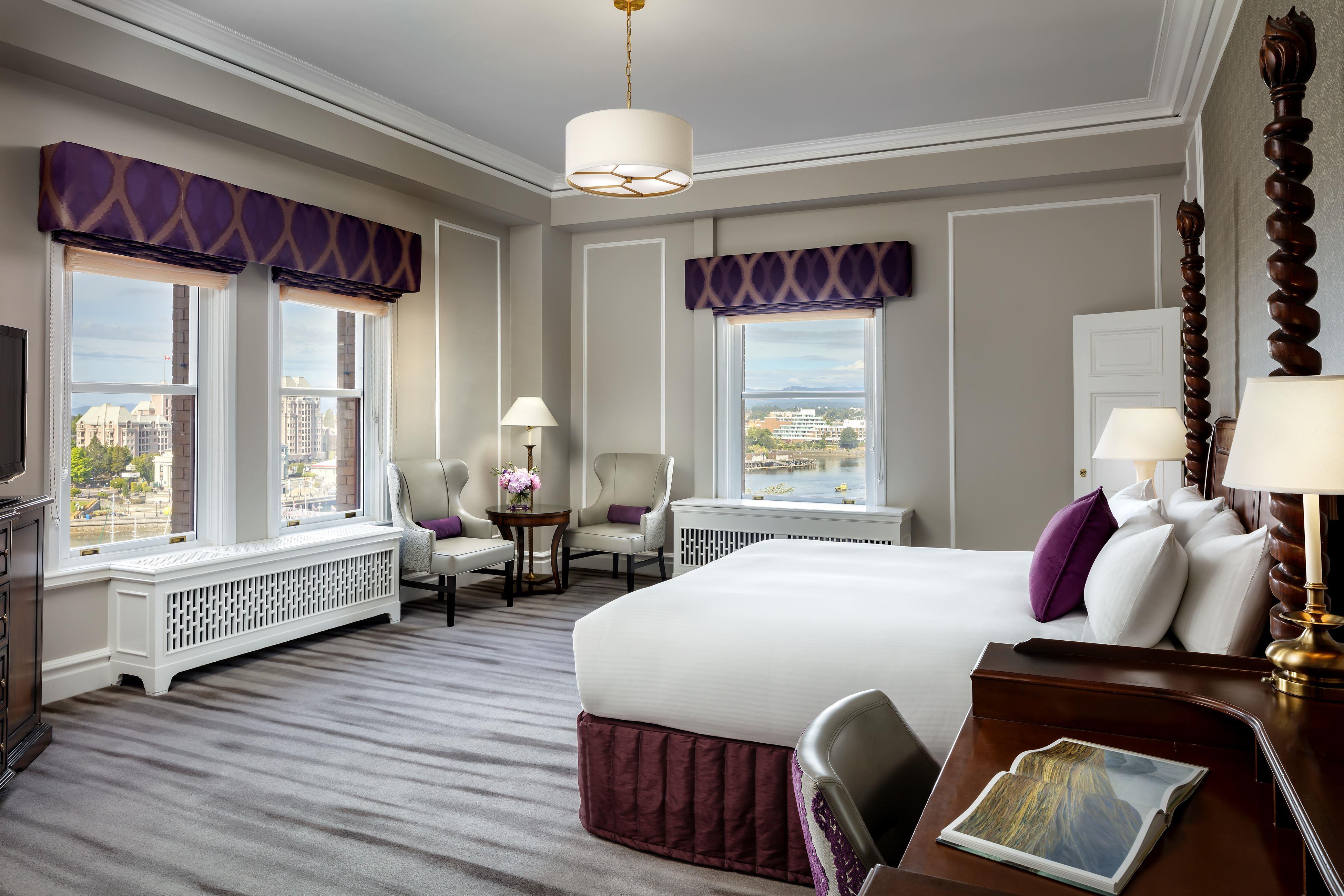 Elegant hotel room featuring a four-poster bed with white linens and purple accents, large windows offering city and harbor views, a seating area with two armchairs, and a writing desk in the foreground.