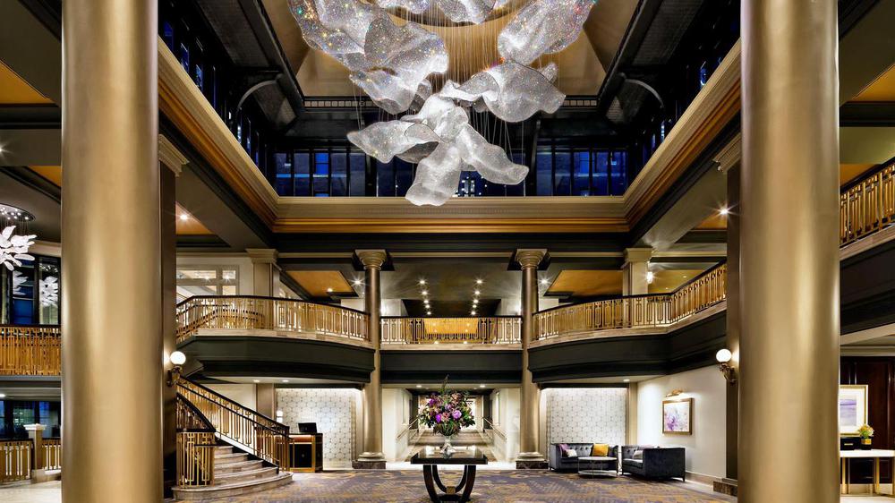 Grand multi-level hotel atrium lobby with gold columns, brass railings, a sweeping staircase, and a dramatic flowing crystal sculpture hanging from the ceiling.