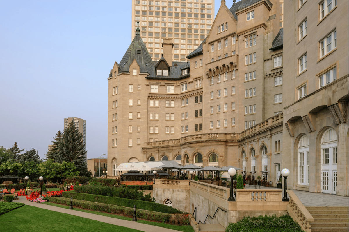 Exterior view of a majestic grand hotel featuring historic château-style stone architecture with turrets and pitched roofs, an outdoor patio terrace with dining tables, and lush green landscaped gardens and walkways.