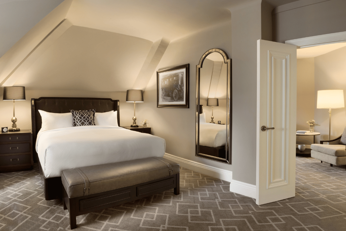 Elegantly styled hotel bedroom under a vaulted ceiling featuring a dark upholstered bed with crisp white linens, a cushioned bench at the foot of the bed, matching nightstands with black lamps, an arched full-length mirror, geometric patterned carpet, and