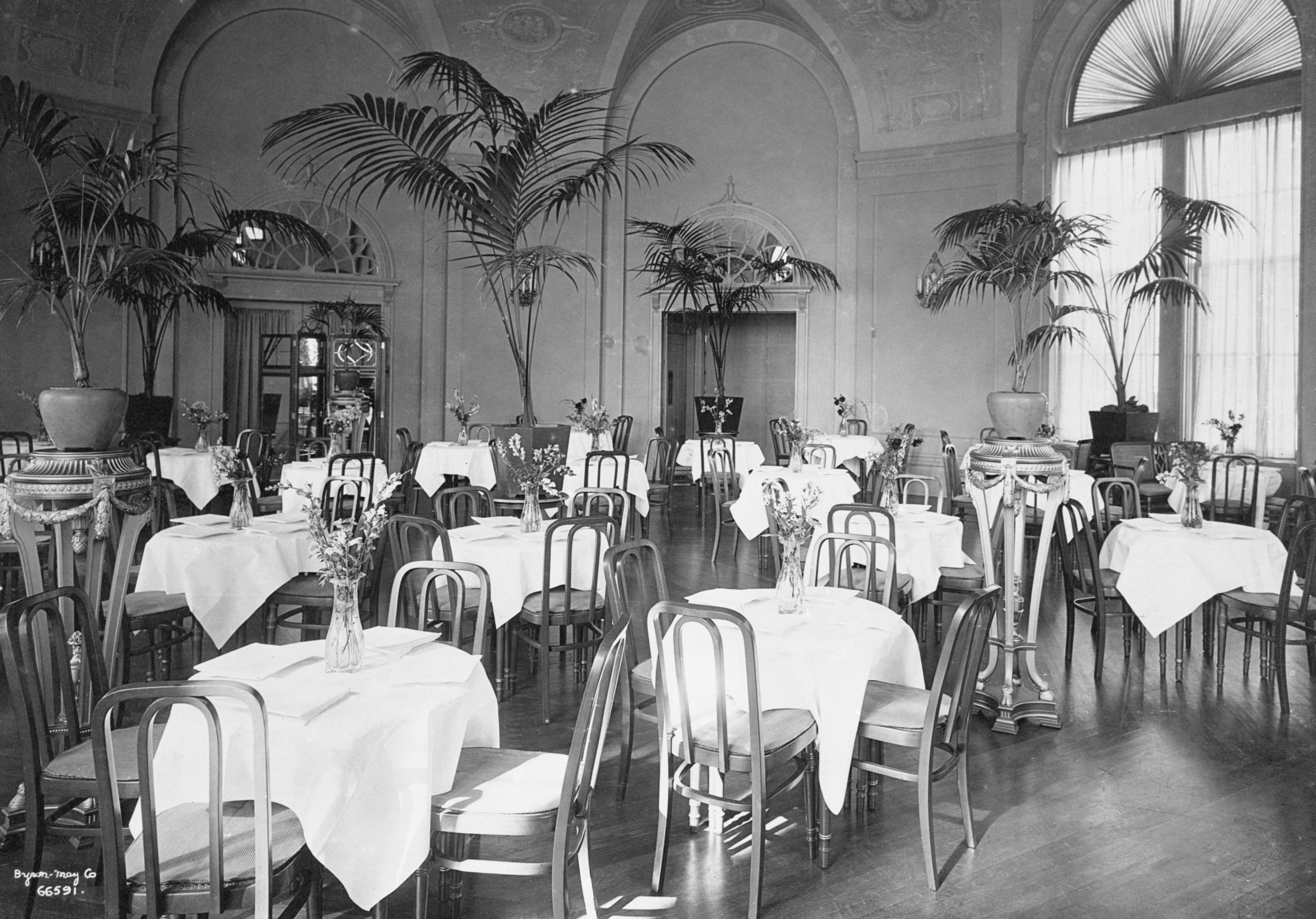 Vintage black-and-white photo of an elegant dining room set with small round tables covered in white tablecloths, bentwood chairs, potted palm trees on decorative pedestals, arched doorways, and large fanlight windows.
