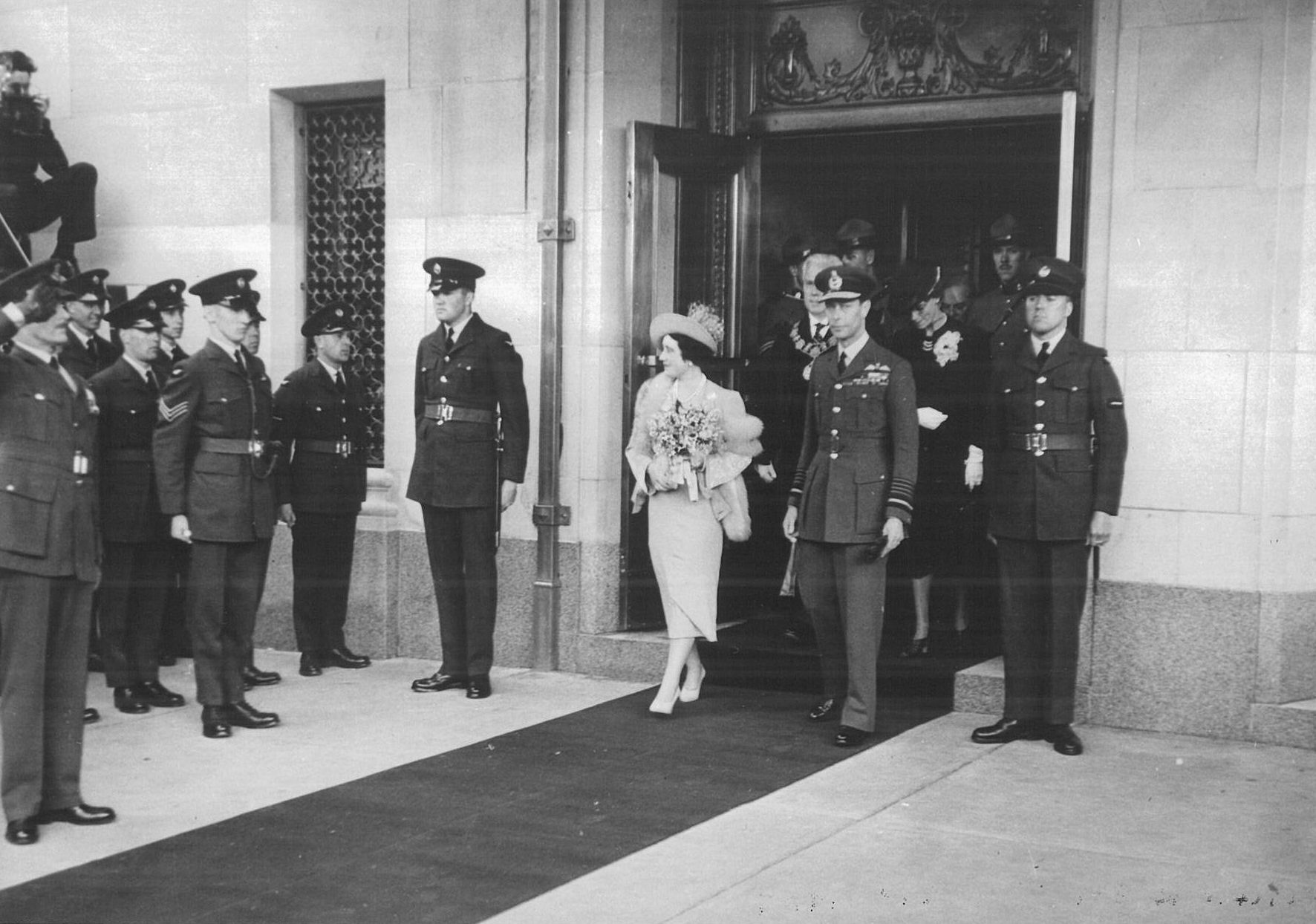 a woman in a dress walking down a sidewalk with a group of people in uniform