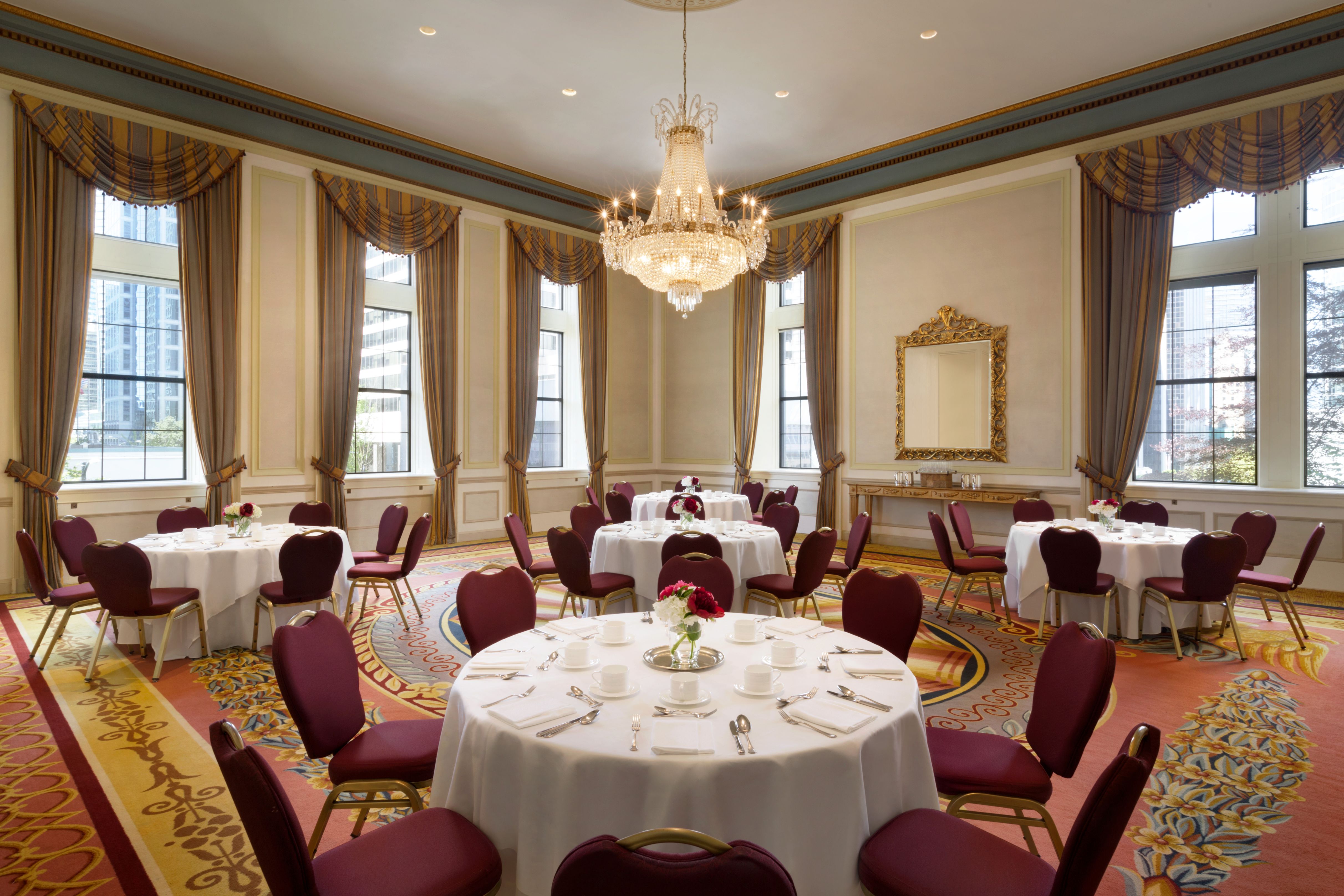 A grand, high-ceilinged ballroom set with round dining tables, white linens, and maroon chairs, featuring tall windows with heavy drapery and a central crystal chandelier.