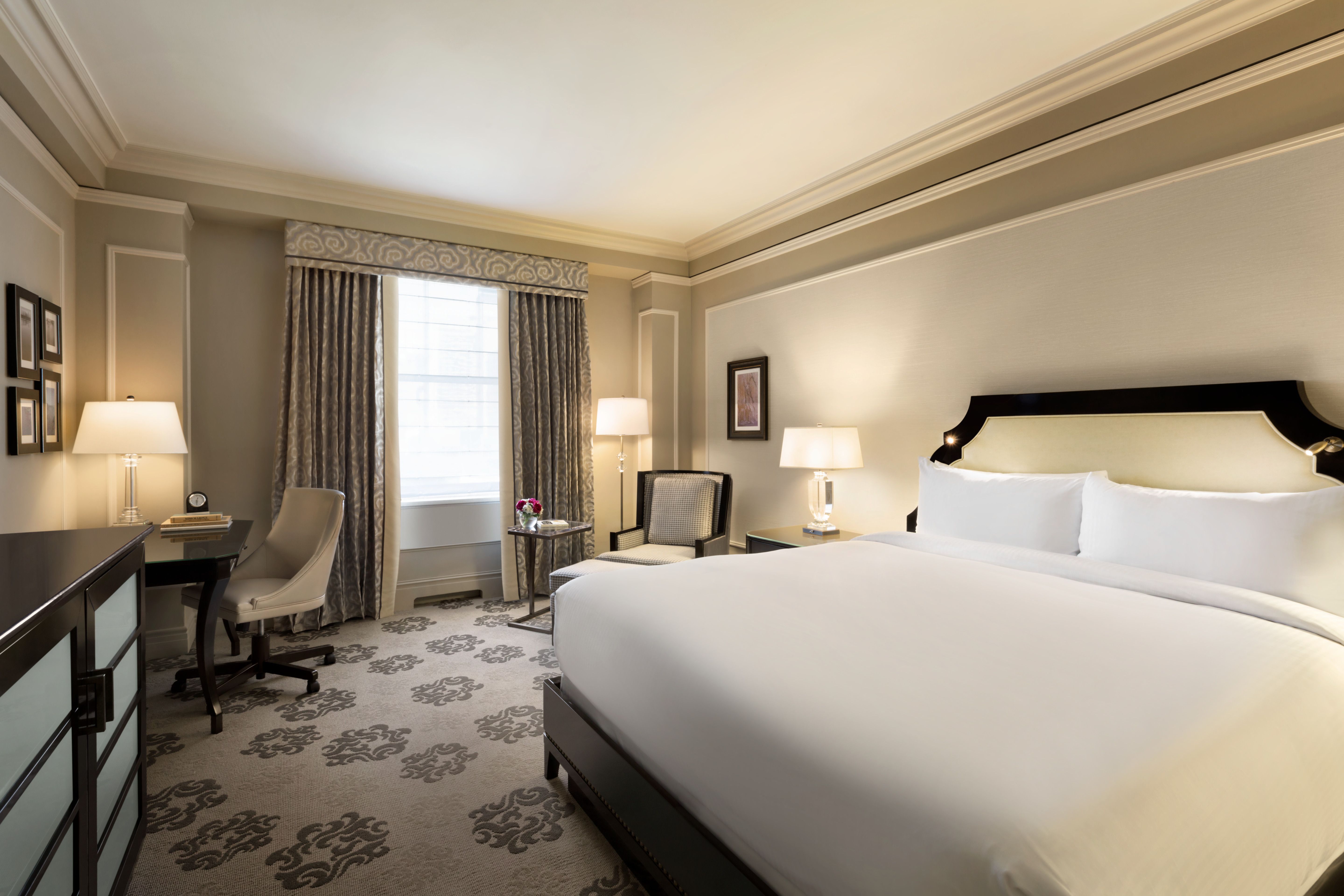 A classic hotel suite featuring a neatly made bed with white linens, a corner armchair with an ottoman, a writing desk with a chair, and patterned grey carpet.