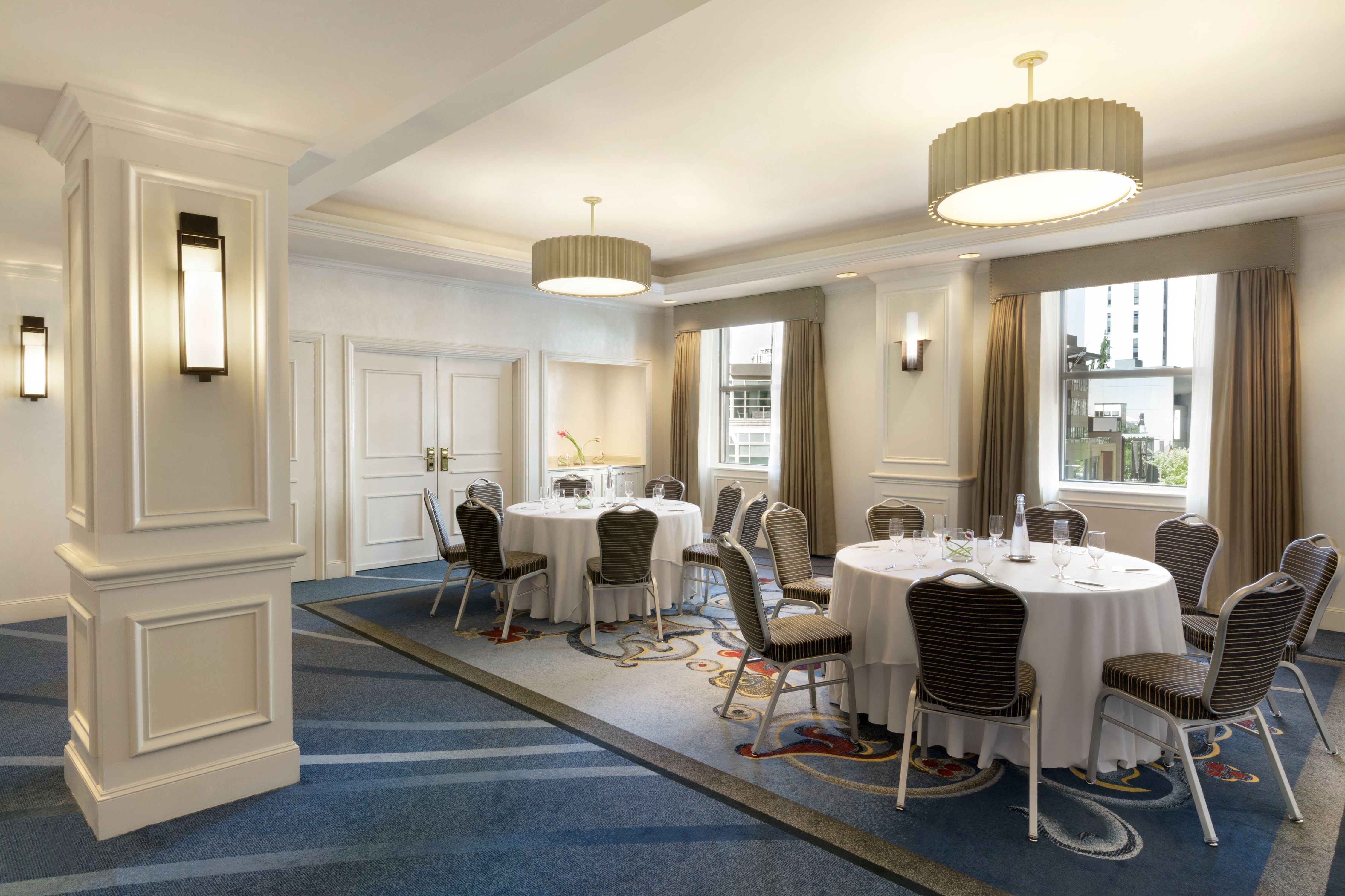 A well-lit hotel meeting room set up with round tables, white tablecloths, dark patterned chairs, large circular light fixtures, and a blue patterned carpet.