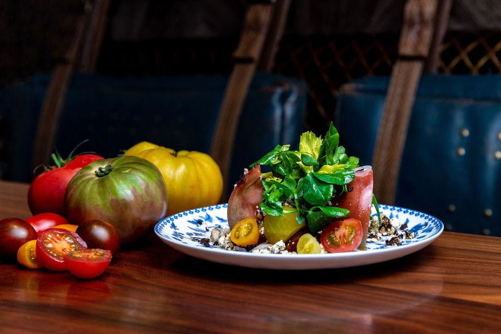 A gourmet tomato salad topped with fresh greens served on a blue and white patterned plate, with whole heirloom tomatoes alongside it on a wooden table.