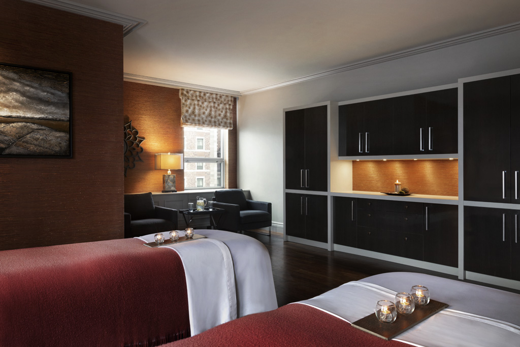 Modern spa treatment room with two massage tables dressed in red and white linens, ambient warm lighting, dark wooden cabinetry, and a cozy seating area near a window.