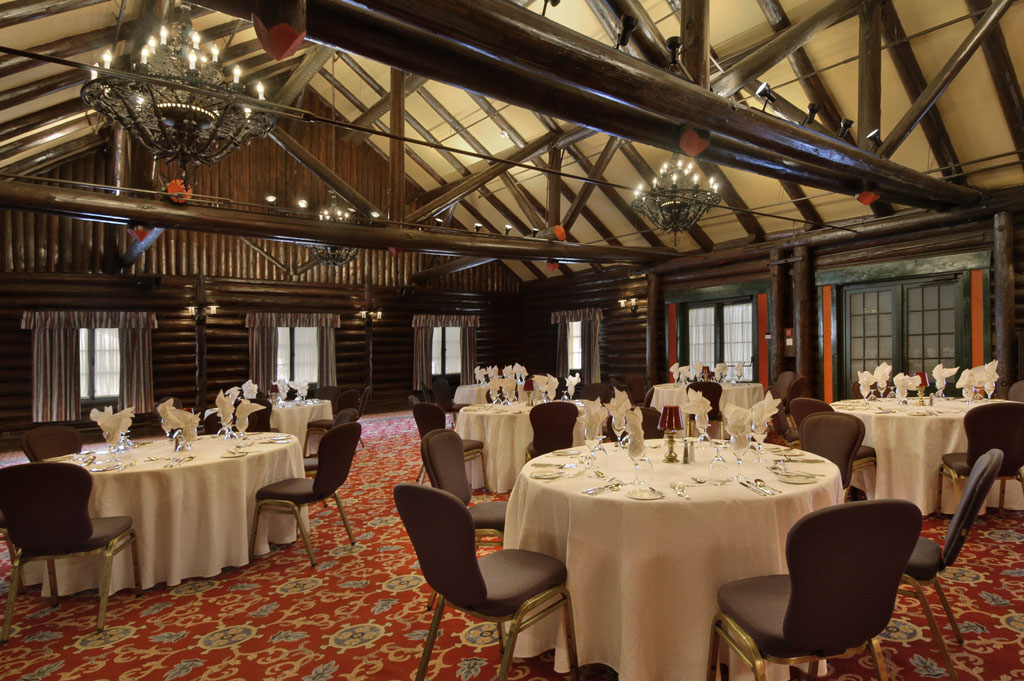 A large rustic banquet room featuring dark log cabin walls, high open-beamed ceilings, wrought-iron chandeliers, and a red patterned carpet. Round event tables are set with white tablecloths, folded napkins, stemware, and dark-padded chairs throughout the