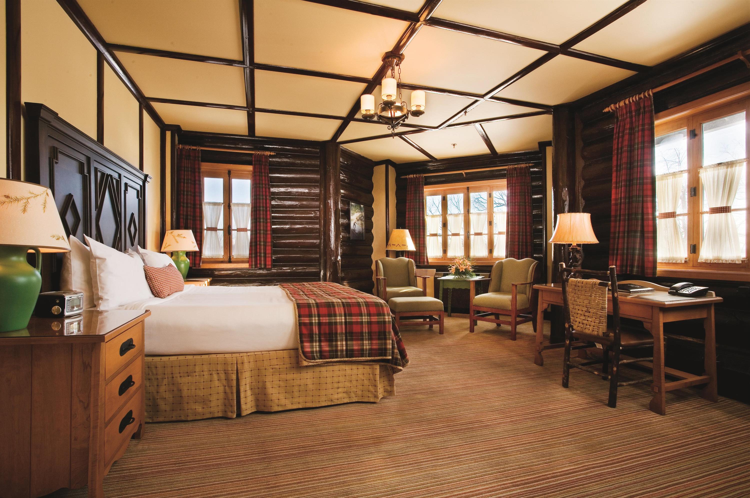 A rustic cabin-style hotel bedroom featuring dark log walls, coffered wooden ceiling, a king bed with a plaid throw blanket, a small sitting area with green armchairs near windows with plaid curtains, and a wooden writing desk with a woven chair.