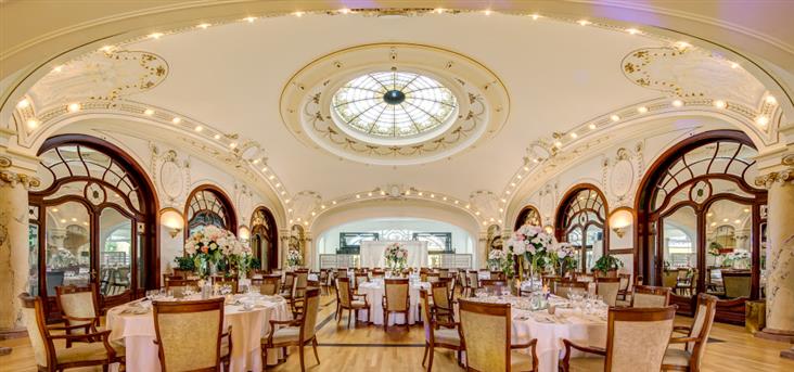 Grand, ornate hotel banquet hall featuring a central circular glass skylight ceiling, arched wooden doorways, light wooden floors, and round dining tables decorated with tall floral centerpieces and beige-upholstered chairs.