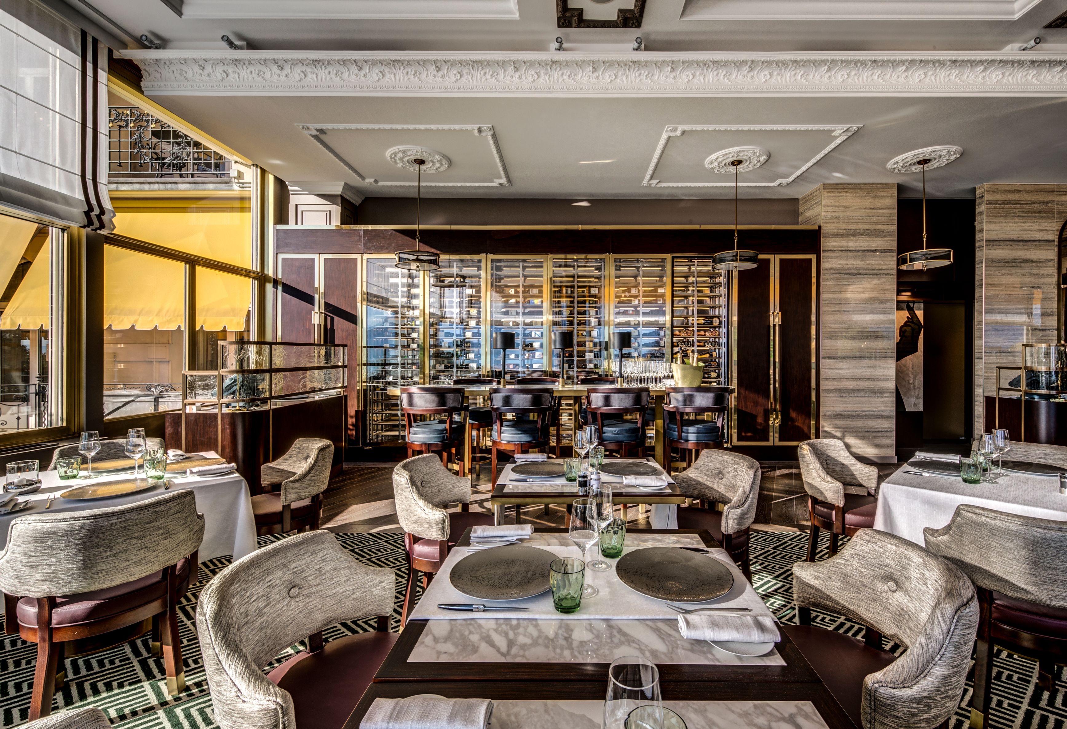 Upscale restaurant dining room featuring geometric patterned carpet, marble-topped tables set with dark plates and green water glasses, plush grey tub chairs, and a large illuminated wine display in the background.