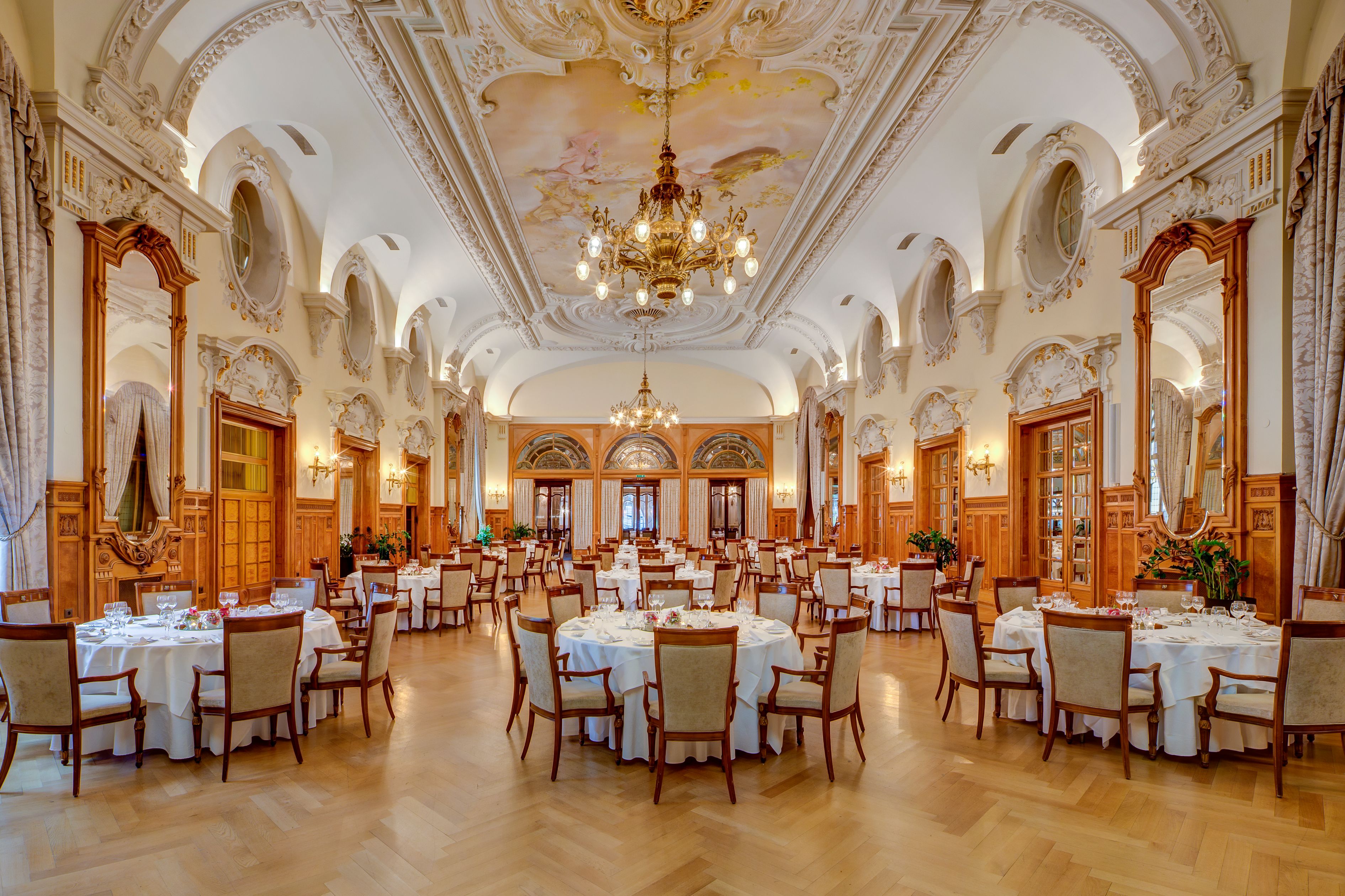 Opulent, high-ceilinged ballroom featuring intricate plaster moulding, ceiling frescoes, grand chandeliers, warm wood wall paneling, herringbone parquet floors, and round dining tables elegantly set with white tablecloths.