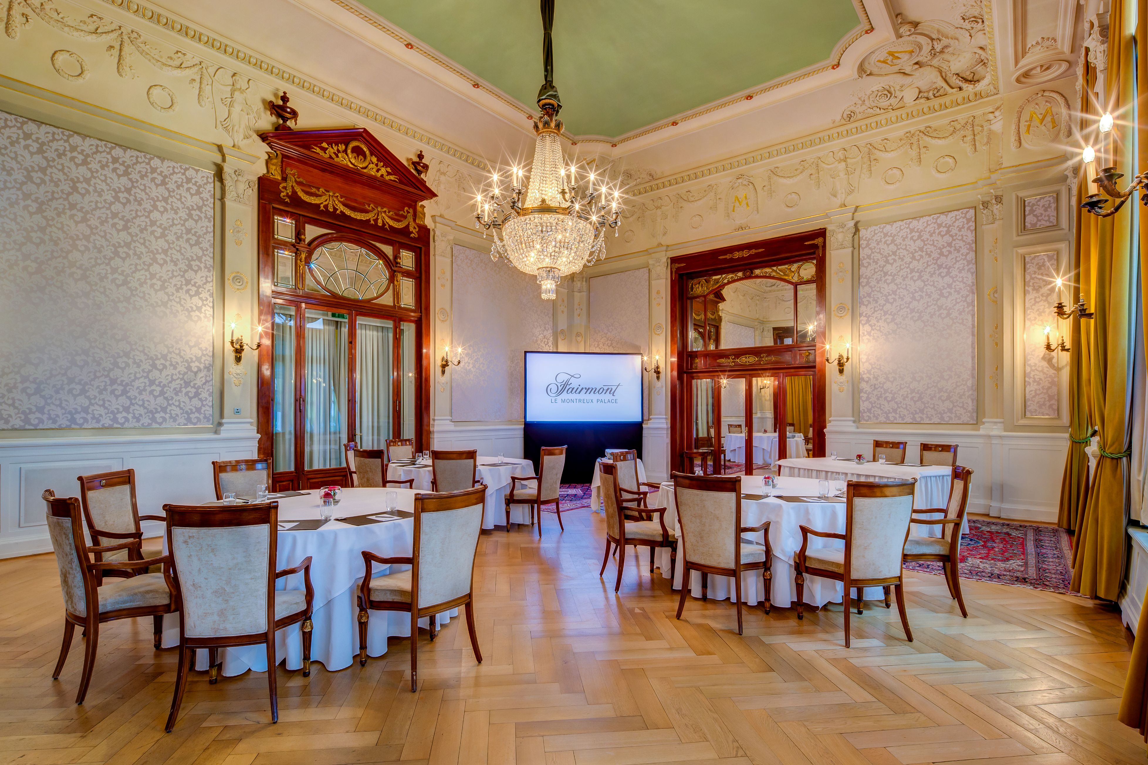 Elegant event hall with a high sage-green ceiling, central crystal chandelier, damask patterned walls, parquet flooring, and round tables prepared for a conference or function.