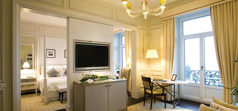 Luxurious, cream-toned hotel suite divided into a cozy bedroom with a white four-poster bed on the left and a bright sitting area with a writing desk and balcony doors on the right.