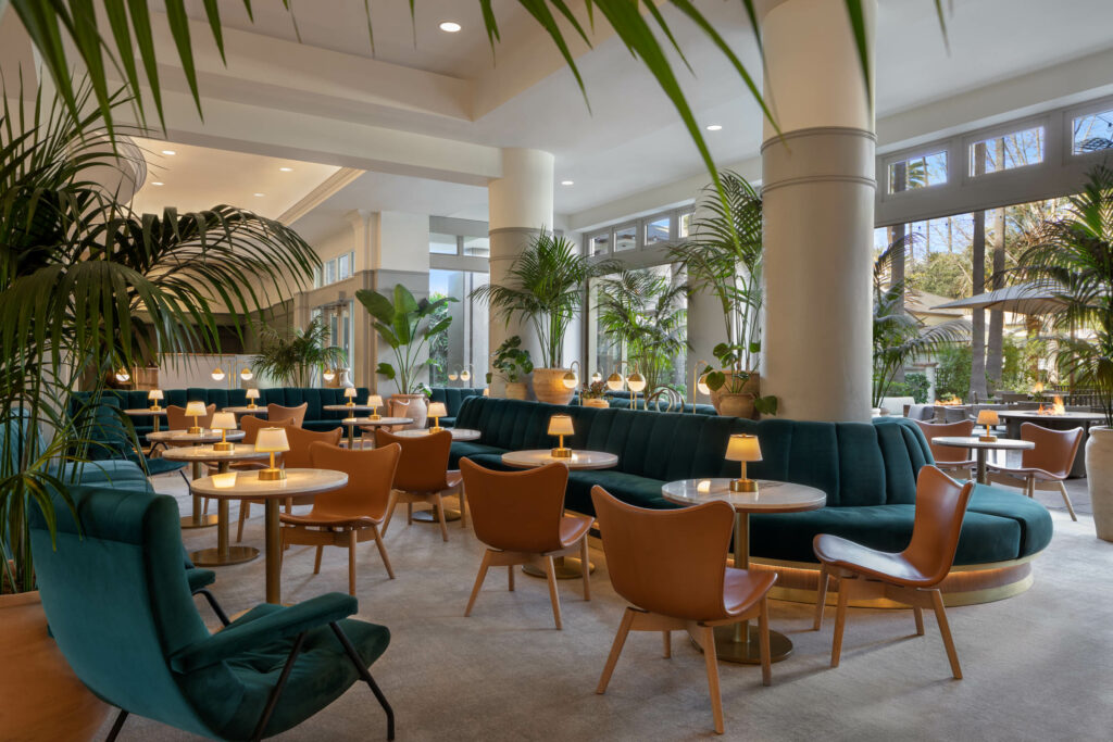 Spacious lounge with large round support pillars, filled with lush tropical potted palms, small white tables lit by brass lamps, warm cognac leather chairs, and long channel-tufted dark teal velvet banquettes.