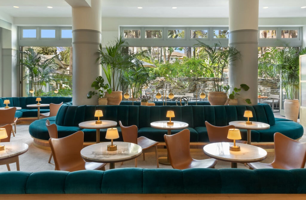 Bright resort lounge with large windows overlooking a lush garden patio, featuring teal velvet channel-tufted seating, small marble-topped bistro tables with warm lamps, leather accent chairs, and large potted palms.