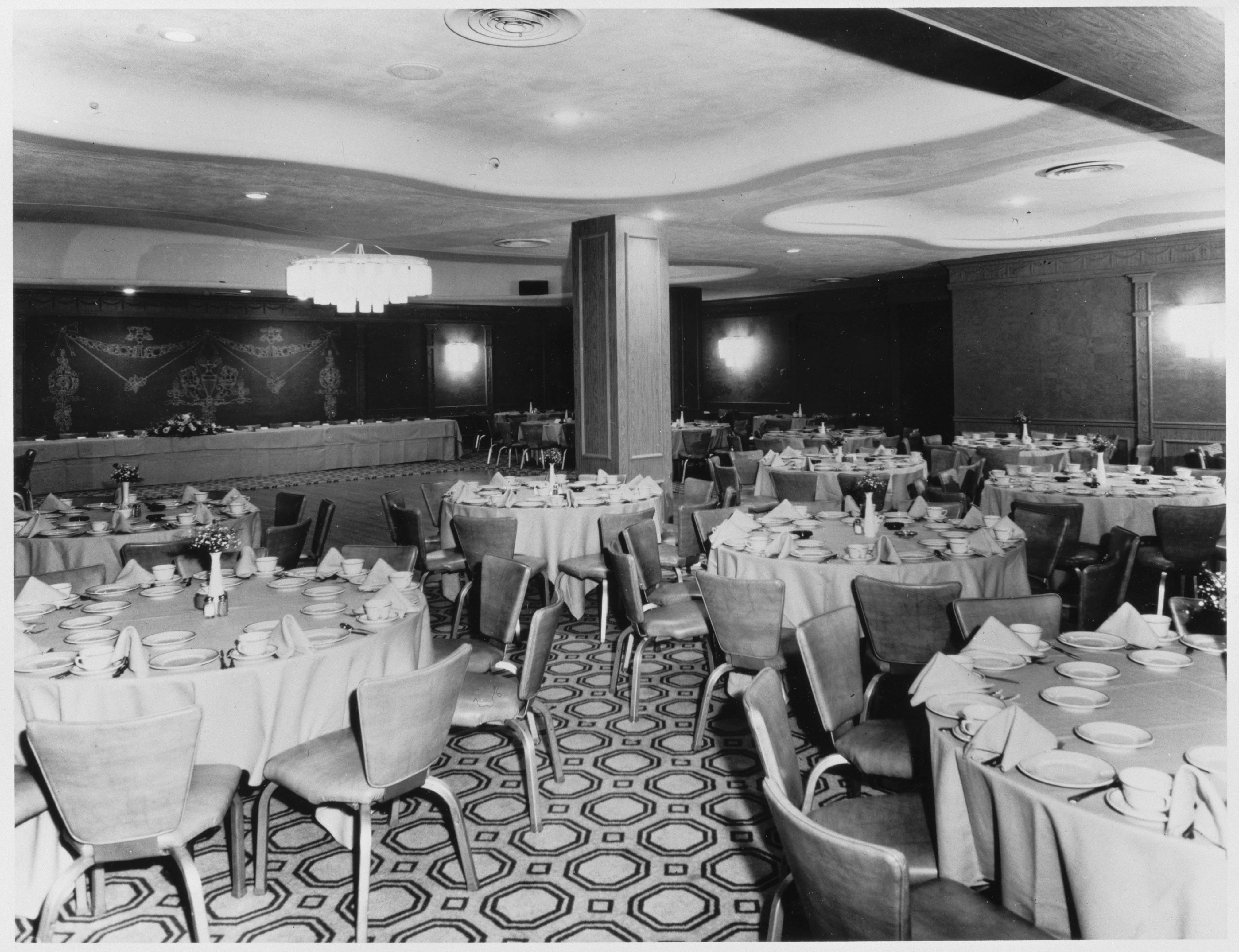 Vintage black-and-white photo of a large banquet hall set up for a formal event with round tables with tablecloths, mid-century style chairs, a geometric patterned carpet, and a long head table at the back.