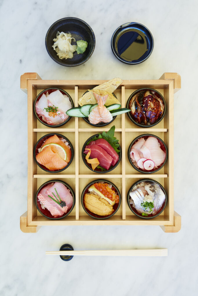 Top-down view of an intricate wooden bento box divided into a nine-grid section, with each compartment holding a small bowl of rice topped with various fresh sashimi and seafood, served alongside soy sauce, wasabi, ginger, and chopsticks on a light marble