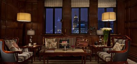 A cozy, upscale hotel lounge featuring rich wood-paneled walls, warm floor lamps, striped armchairs, and a central sofa with decorative throw pillows arranged around a coffee table, set against large windows overlooking a city skyline at dusk.