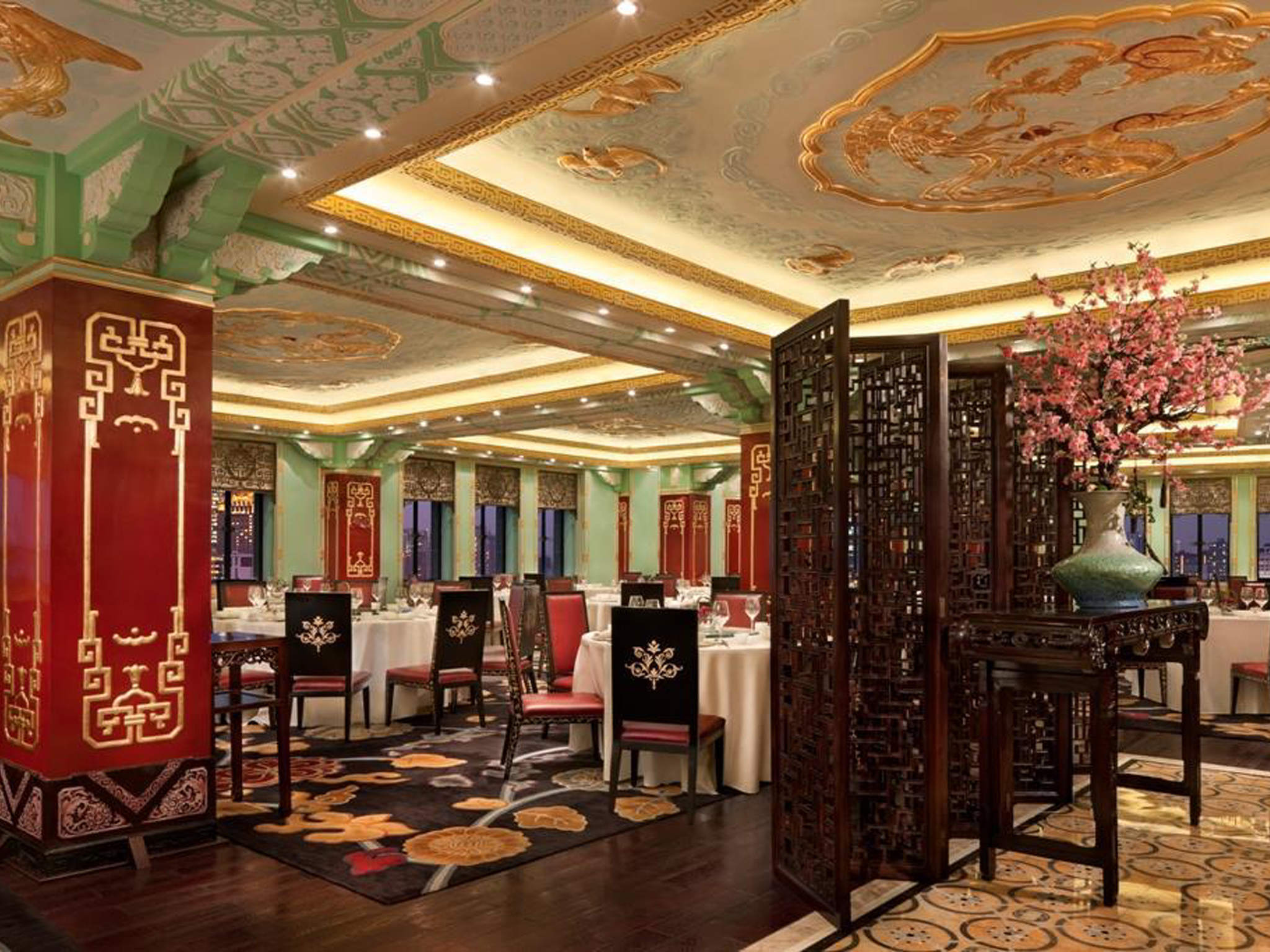 An opulent Chinese fine-dining restaurant interior featuring intricate gold ceiling carvings, red and gold accent pillars, dark wooden dining chairs around white-clothed tables, and a wooden folding screen beside a vase with pink cherry blossoms.