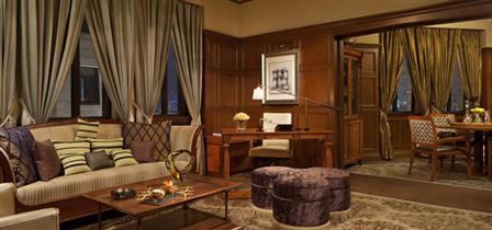An elegant, wood-paneled hotel suite featuring a beige striped sofa with patterned throw pillows, three round purple ottomans around a coffee table, a dedicated desk study area, and an adjoining dining space visible through a wide doorway.