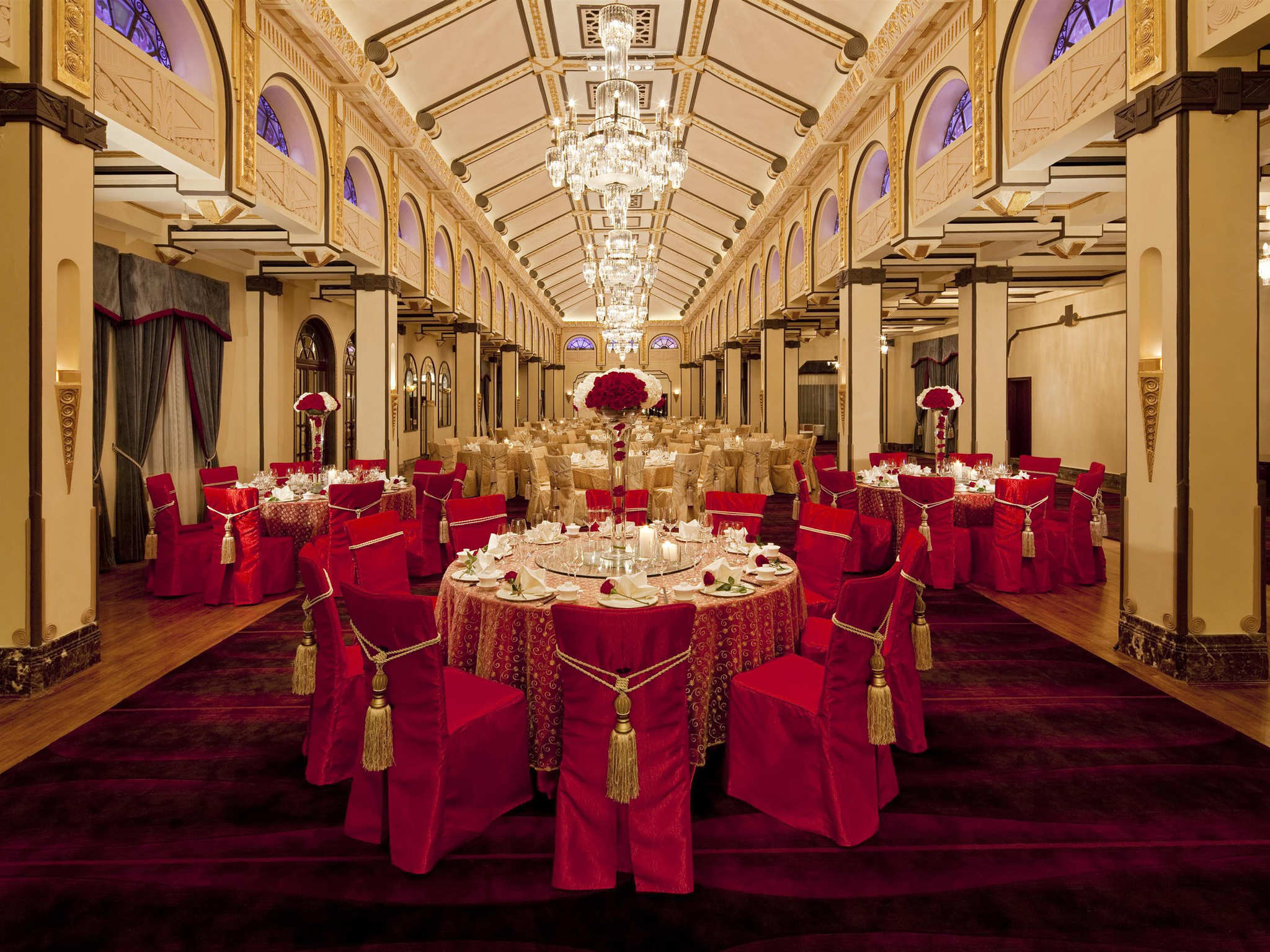 A grand, high-ceilinged hotel banquet hall decorated for an event with crystal chandeliers, vaulted ceilings, and round dining tables elegantly set with red chair covers, gold tassels, and floral centerpieces.