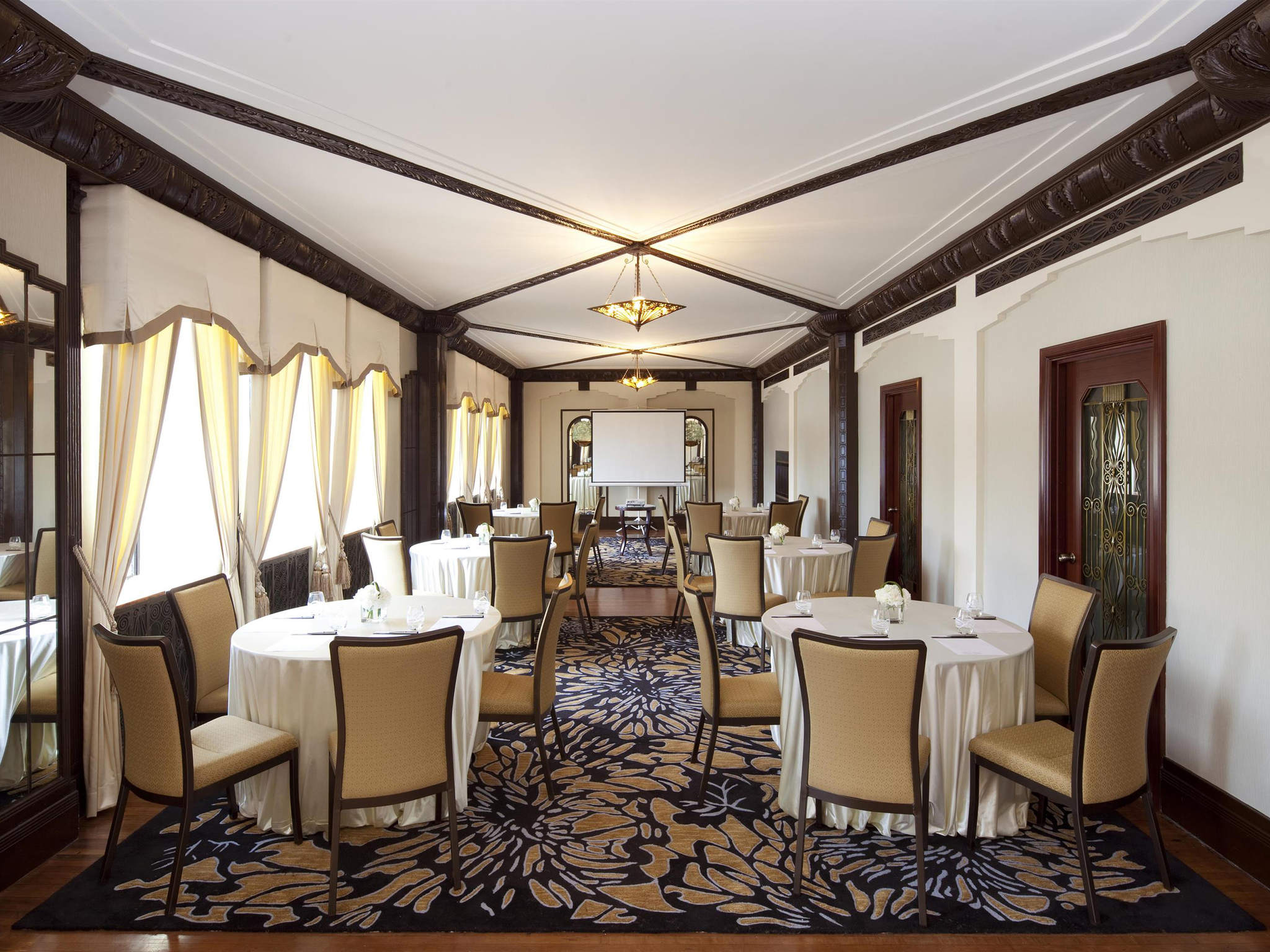 An elegant hotel meeting or event room configured with round tables and beige upholstered chairs set on a patterned carpet, framed by dark carved wood ceiling beams, cream drapery, and a presentation screen at the far end.