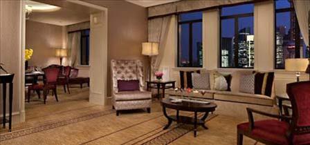 A luxurious hotel suite living area featuring a neutral sofa with decorative pillows along a row of windows overlooking a night skyline, accompanied by an upholstered wingback armchair, a dark wood coffee table, and an adjoining dining room.